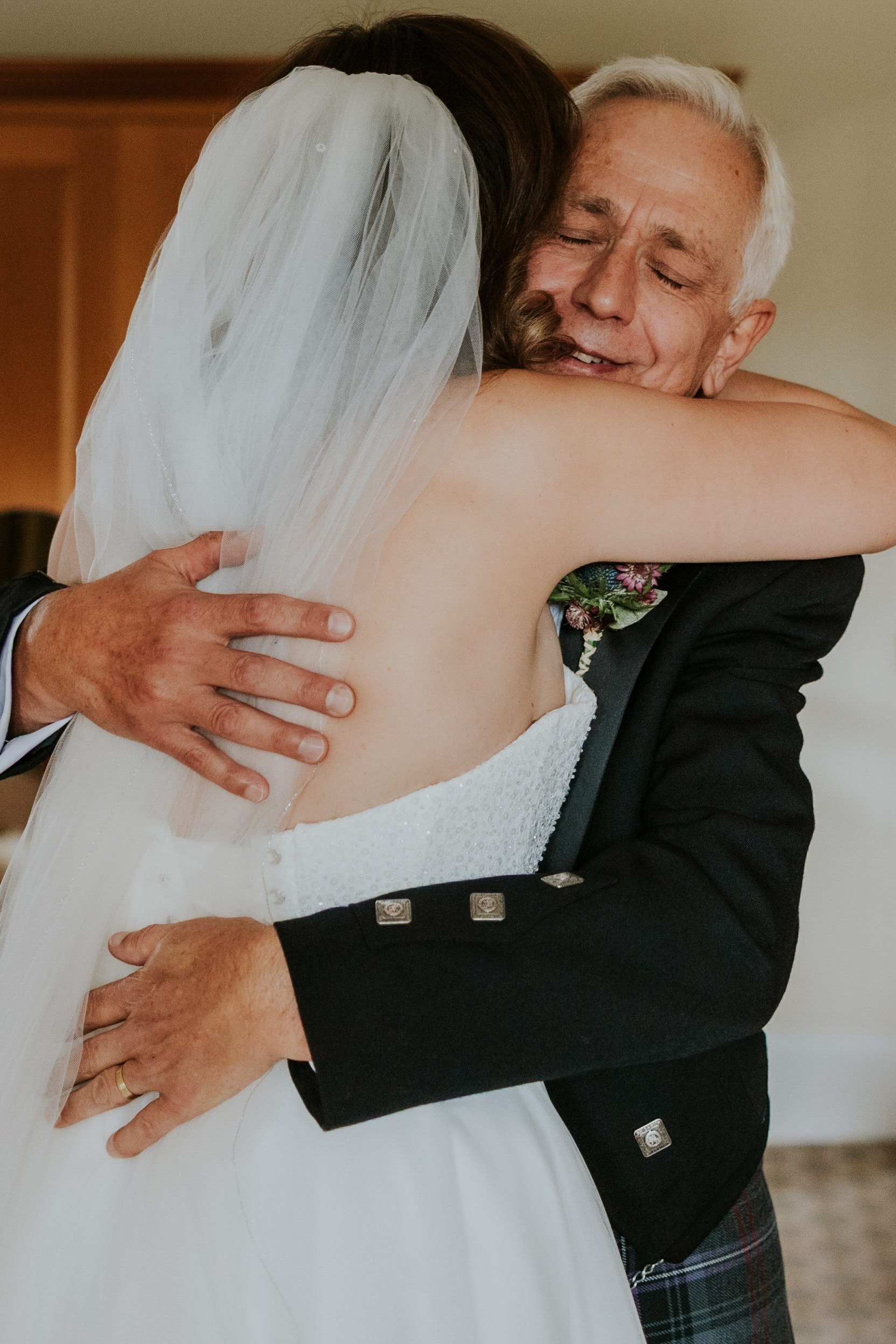 Bride in a white dress and veil hugging a grey-haired man in a black jacket and tartan kilt.