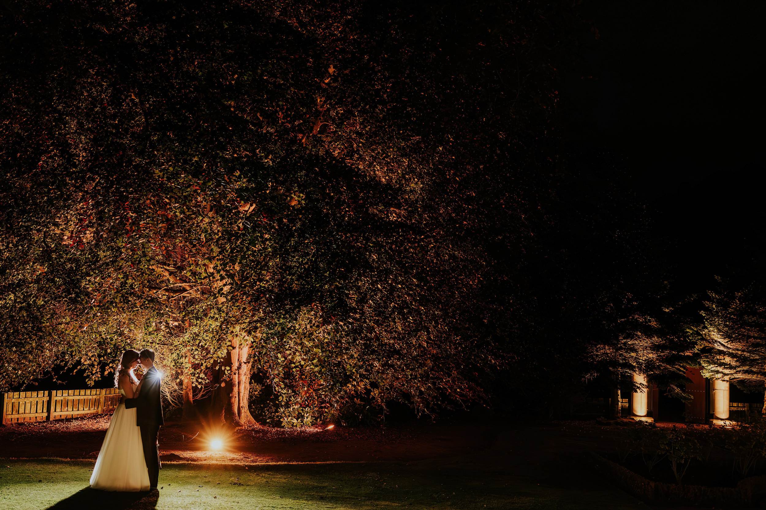 Emma and Gordon embracing beneath illuminated trees on the lawn at Lochgreen House Hotel at night.