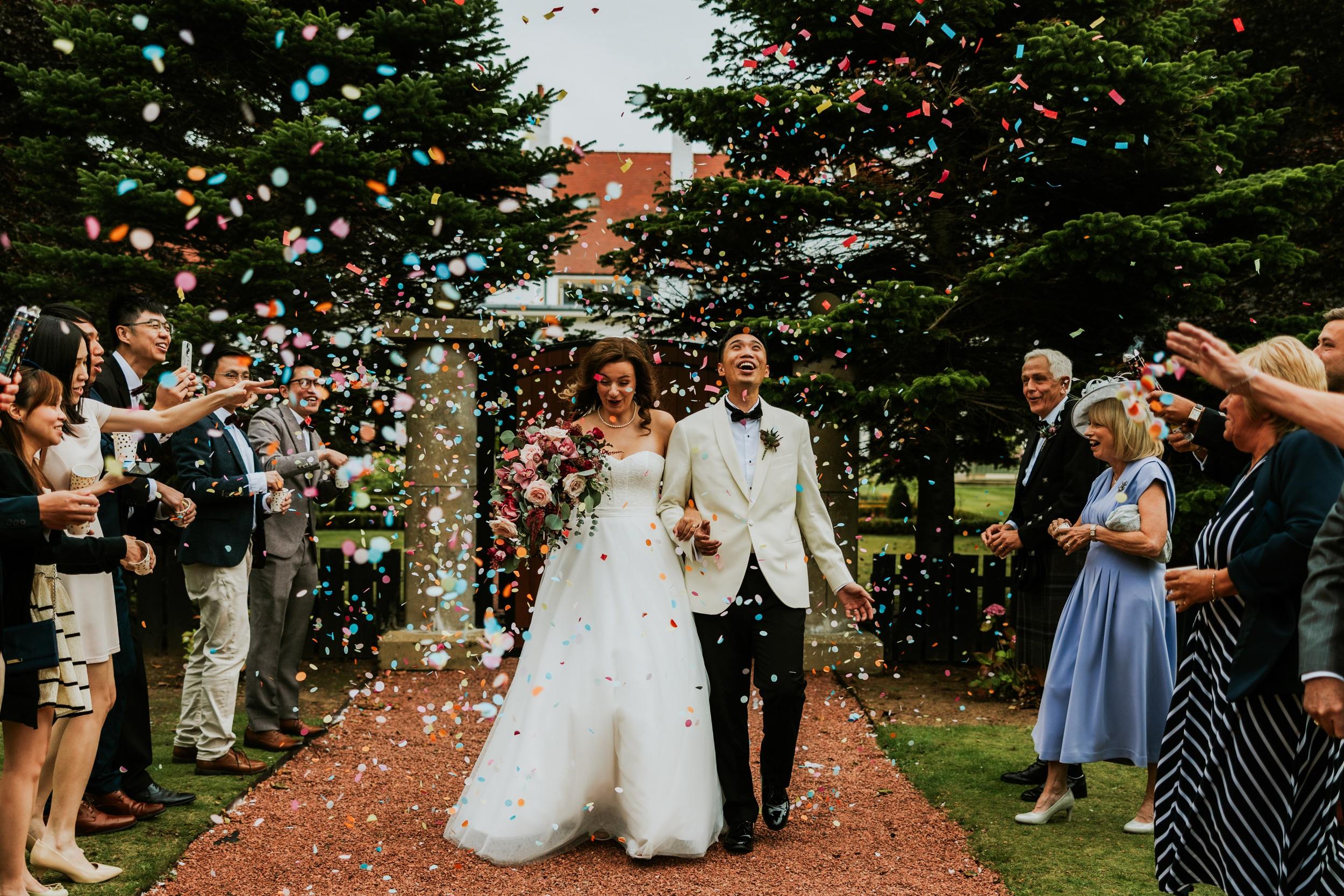 Emma and Gordon holding hands as guests shower them with colourful confetti at Lochgreen House Hotel.