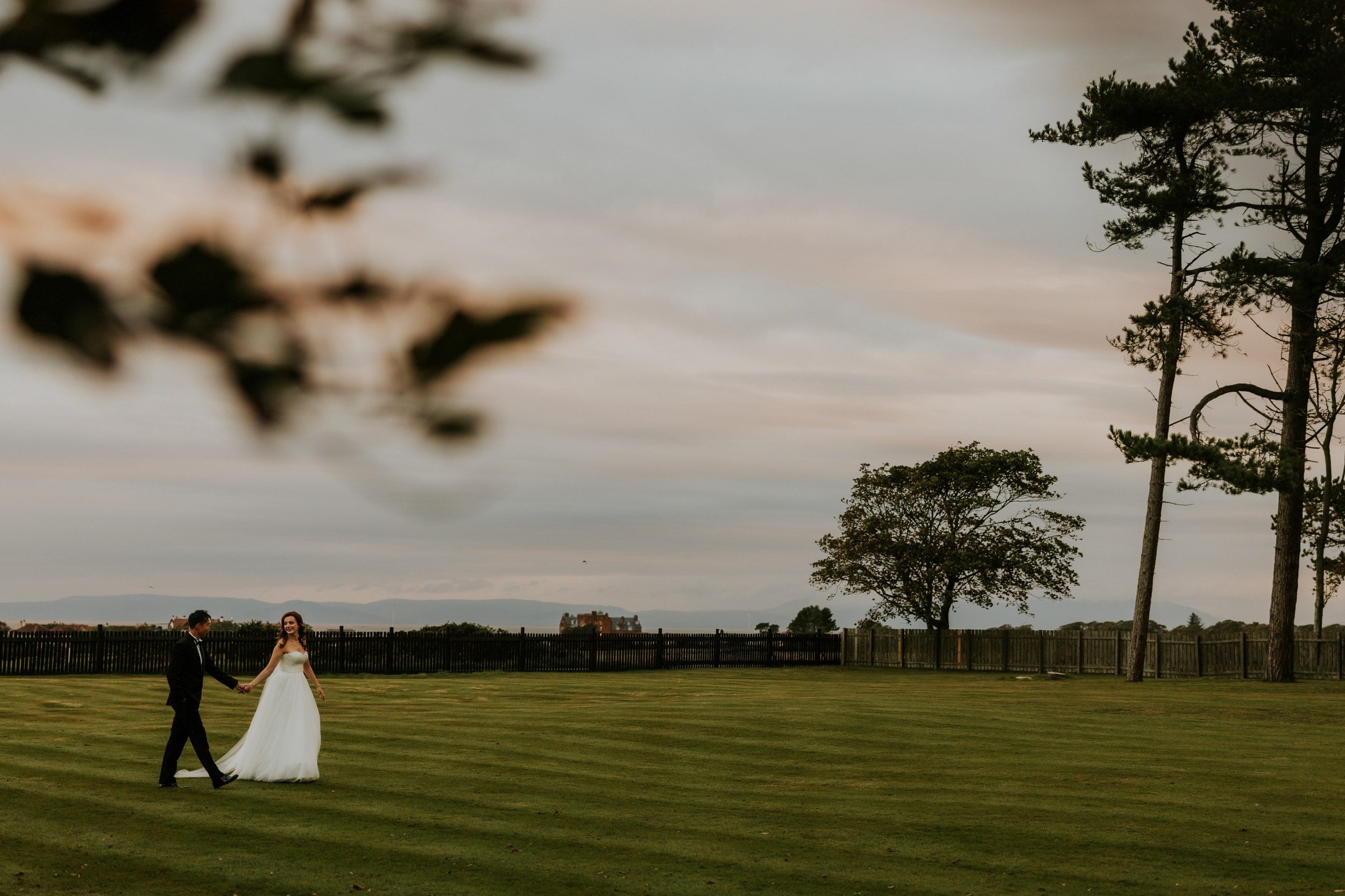 Emma and Gordon holding hands as they walk across the lawn, with tall trees and distant hills behind them.