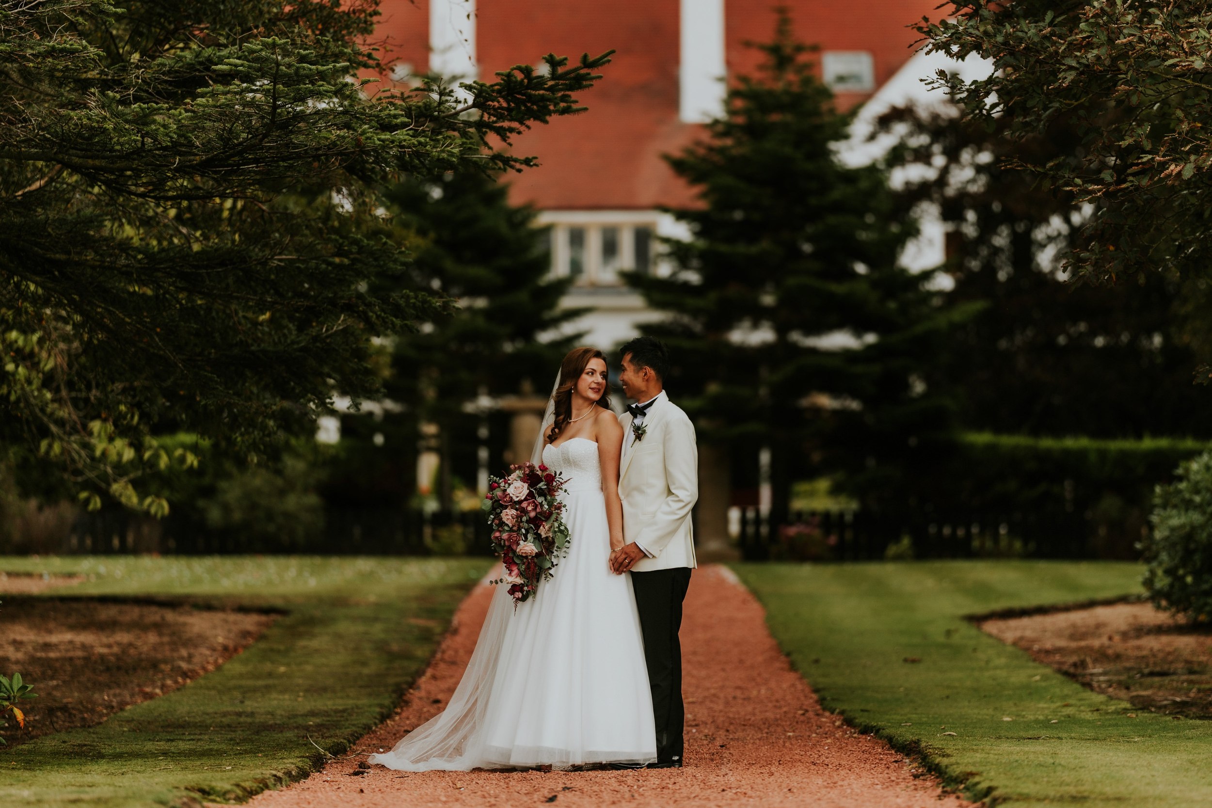 Emma and Gordon holding hands on a tree-lined garden path at Lochgreen House Hotel, with Emma carrying a cascading bouquet.