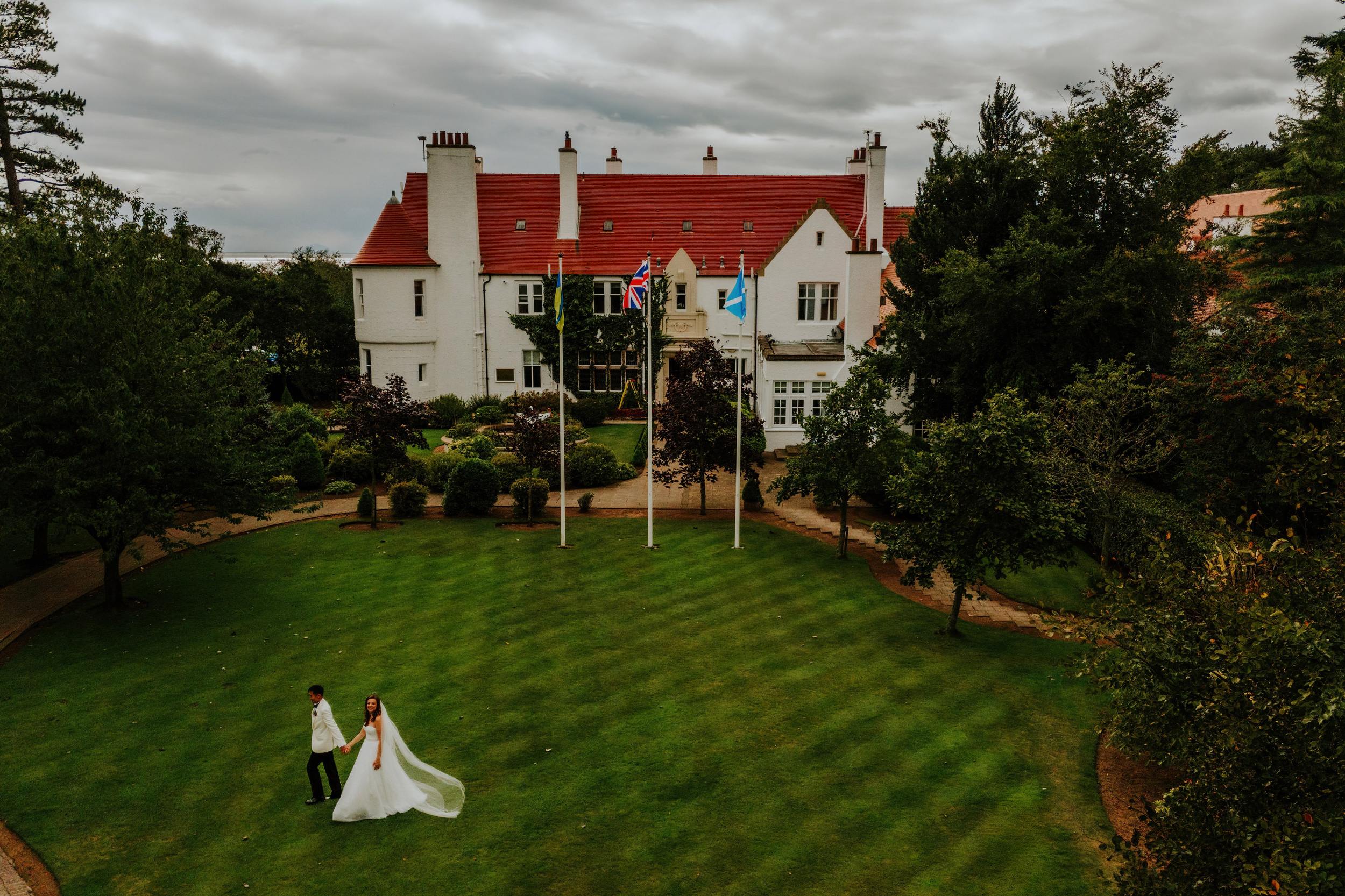 Emma and Gordon holding hands on the lawn in front of Lochgreen House Hotel, with its white walls and red roof behind them.