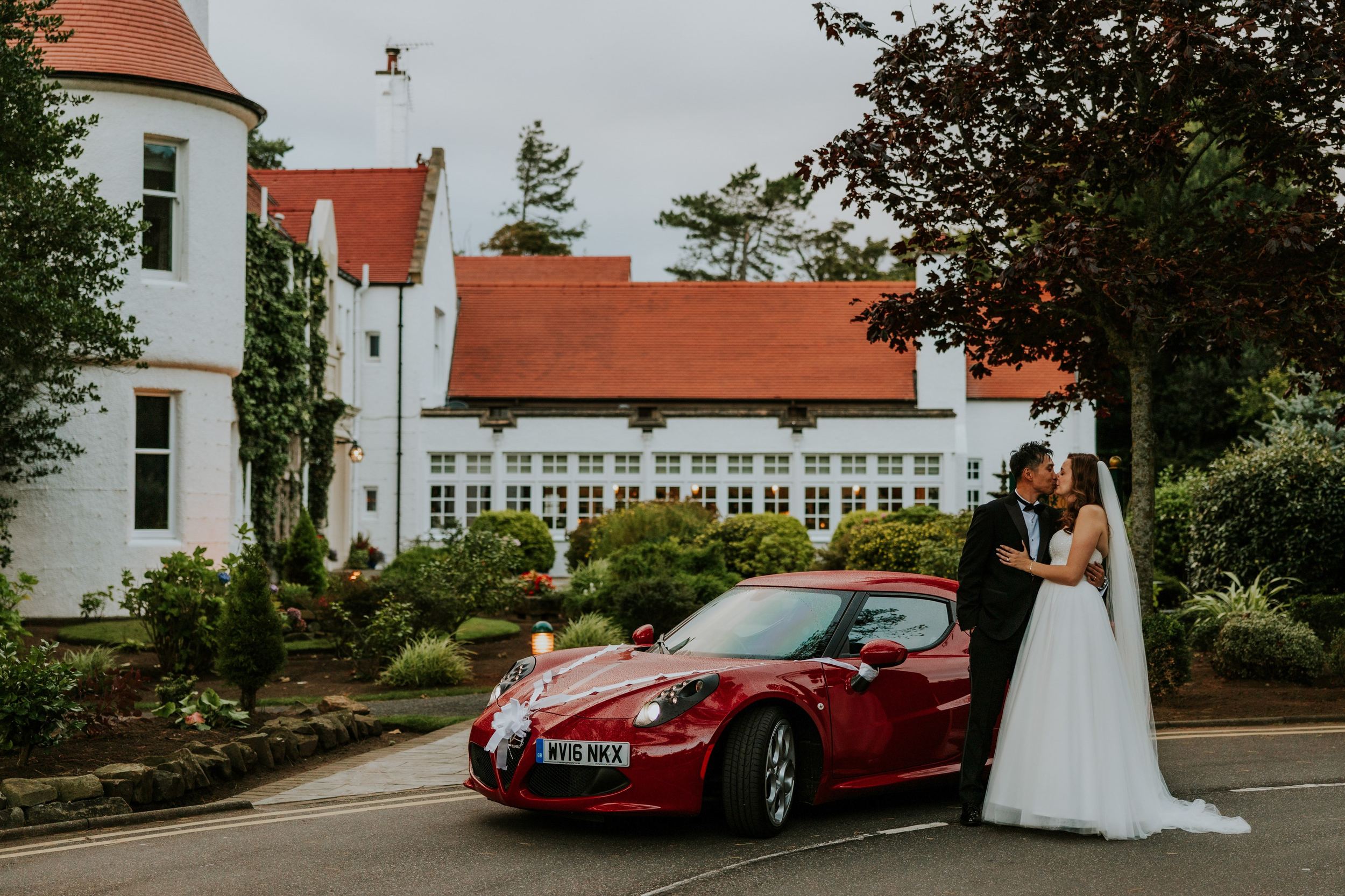 Emma and Gordon kissing beside a red wedding car decorated with white ribbons outside Lochgreen House Hotel.