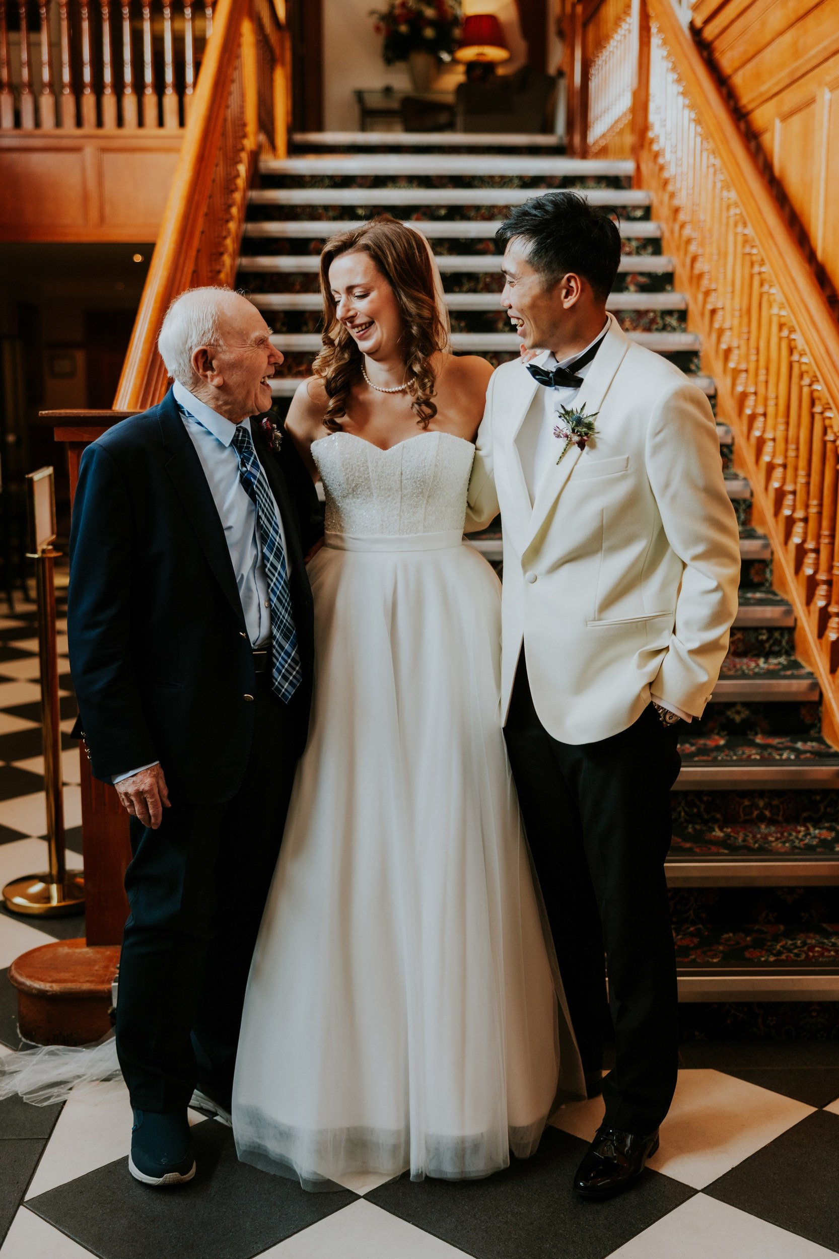 Emma and Gordon laughing with an older guest beside the wooden staircase at Lochgreen House Hotel.