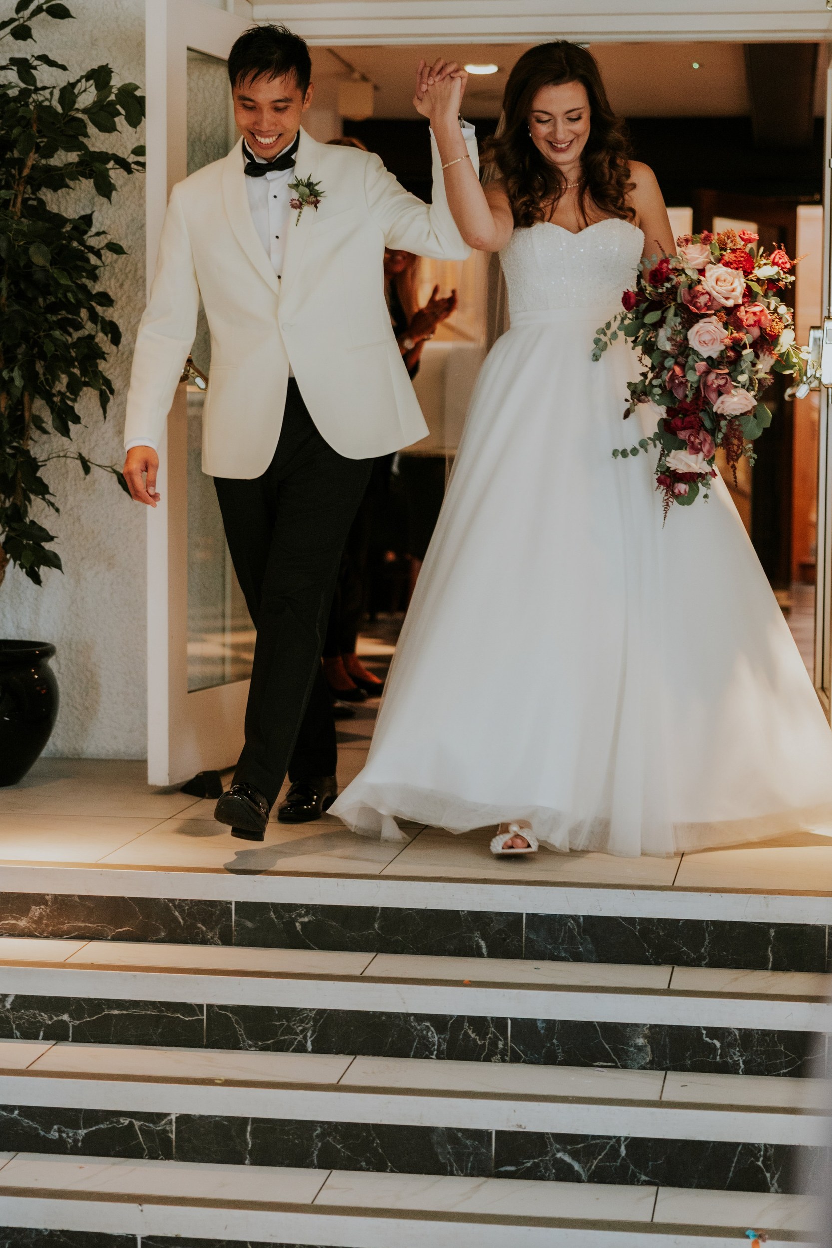 Emma and Gordon raising their joined hands as they approach steps at Lochgreen House Hotel, Emma carrying a cascading bouquet.