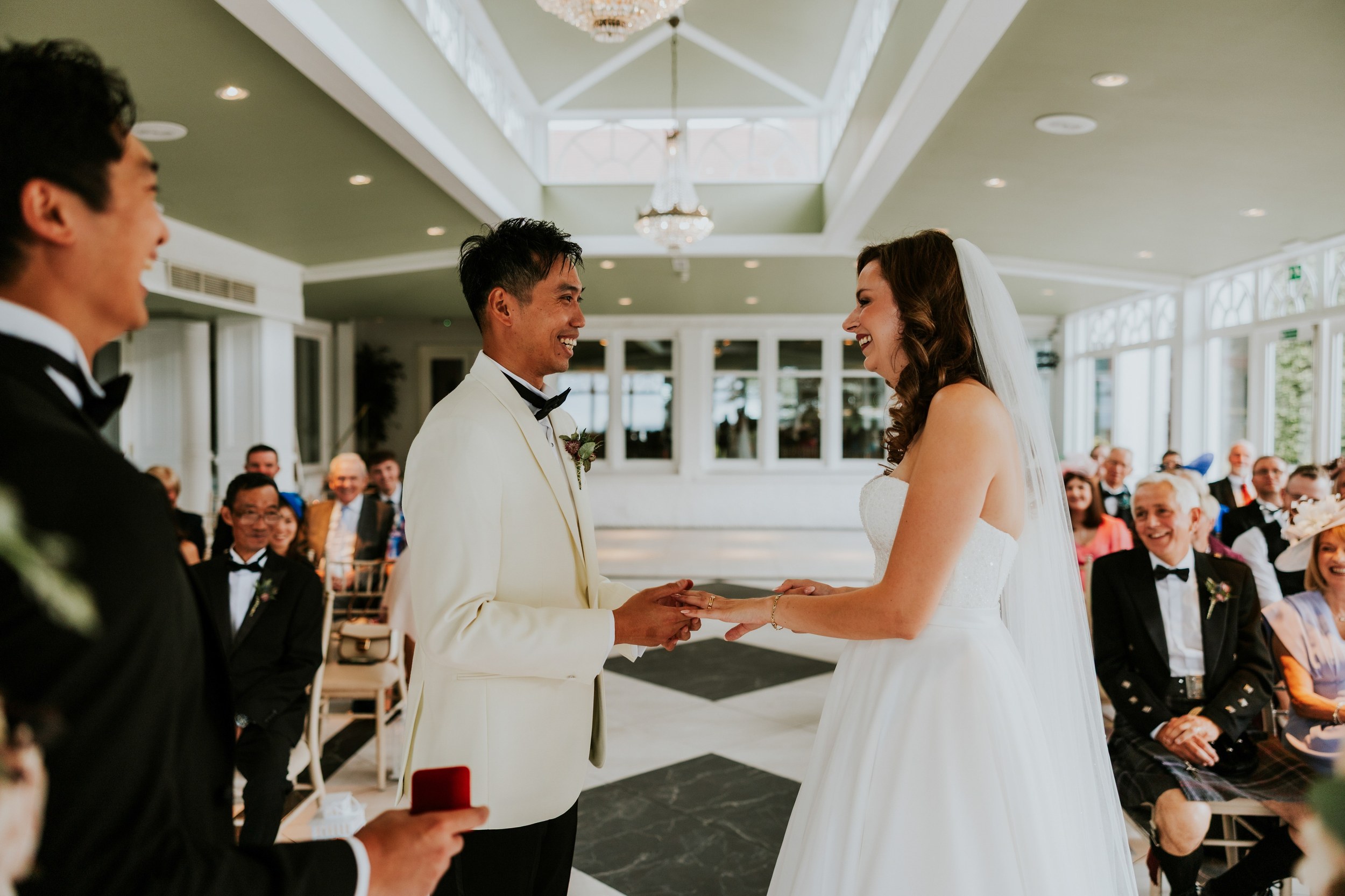 Emma and Gordon smiling and holding hands during their wedding ceremony in the orangery at Lochgreen House Hotel.