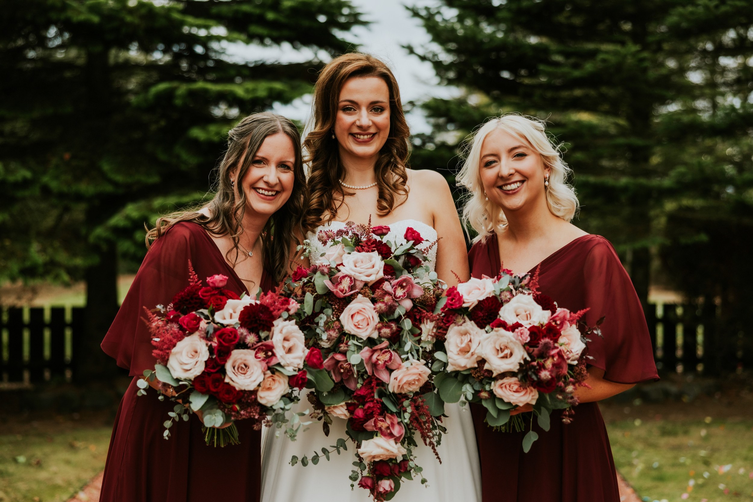 Emma and two bridesmaids in burgundy dresses holding bouquets outdoors at Lochgreen House Hotel.