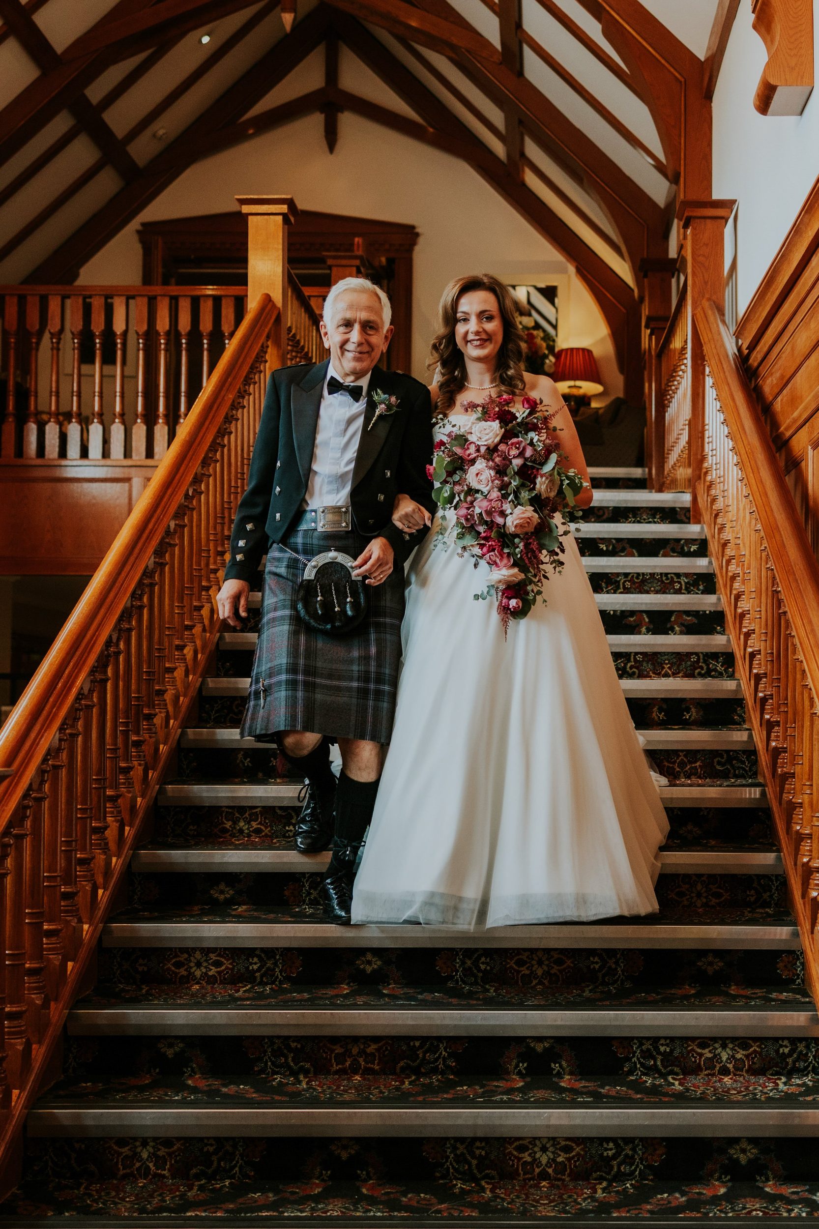 Emma holding her bouquet beside a man in Highland dress on the wooden staircase at Lochgreen House Hotel.