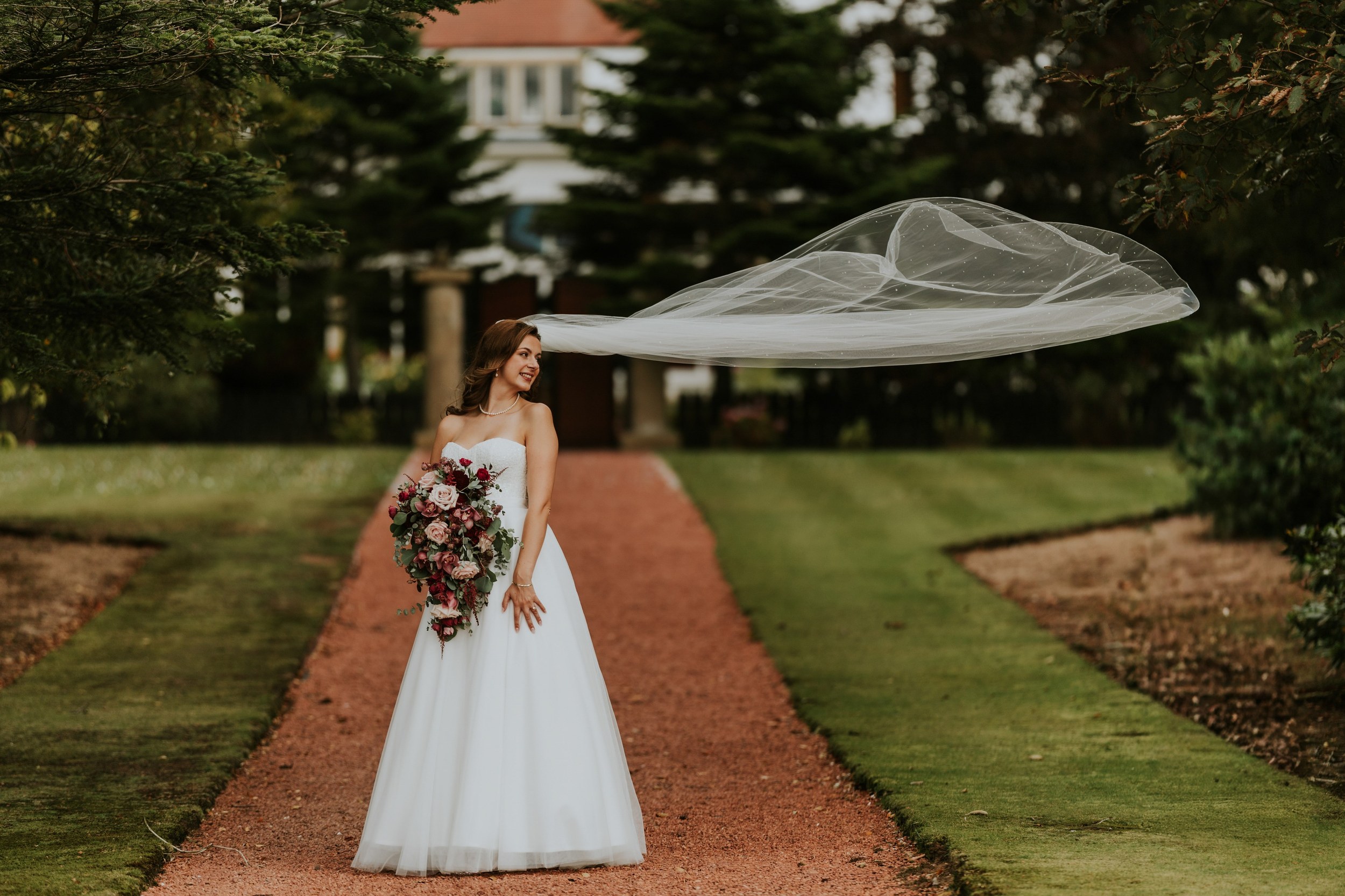 Emma holding her bouquet on a garden path at Lochgreen House Hotel, with her long veil floating to one side.