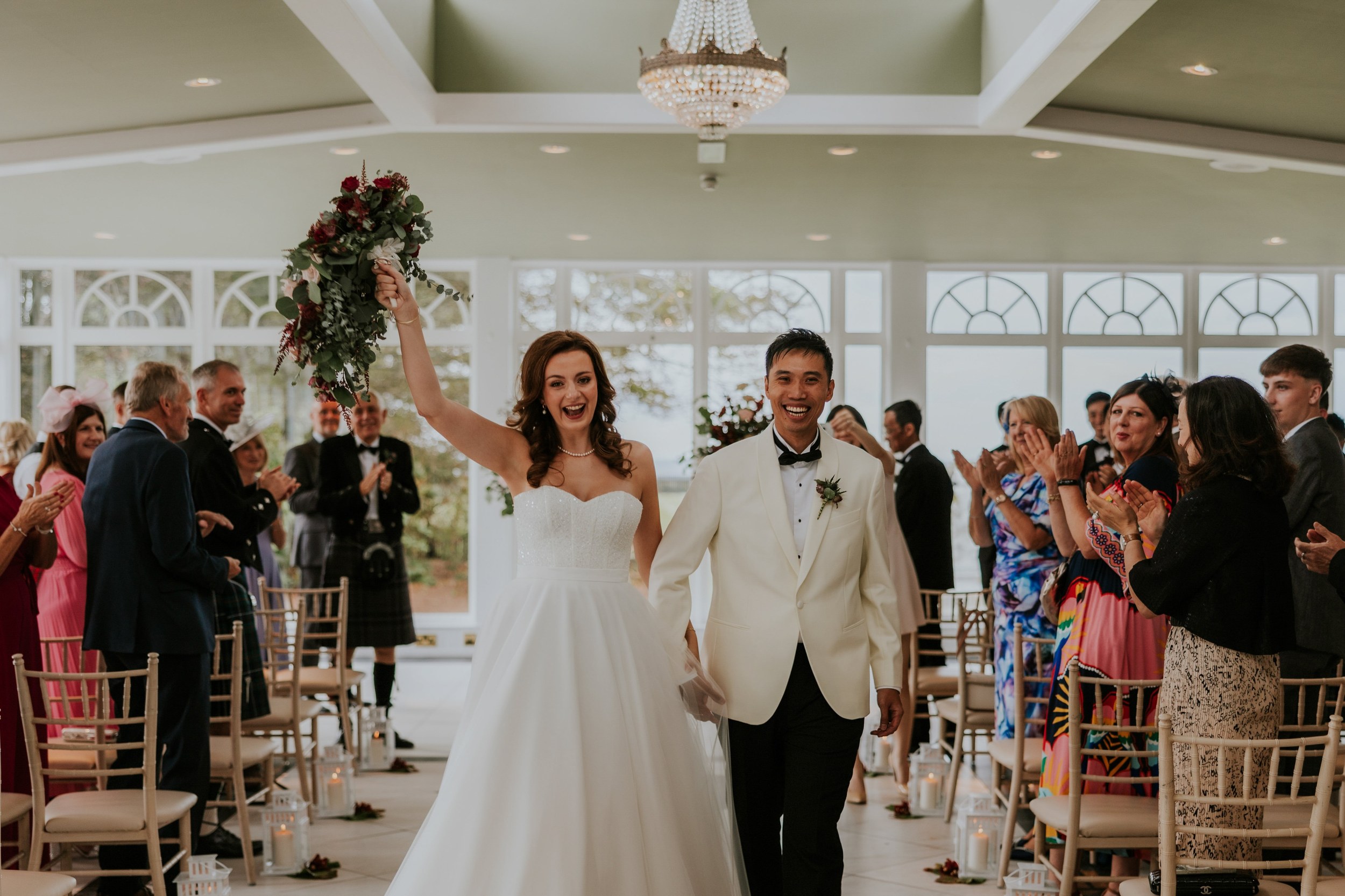 Emma raises her bouquet as she and Gordon walk down the aisle past applauding guests at Lochgreen House Hotel.