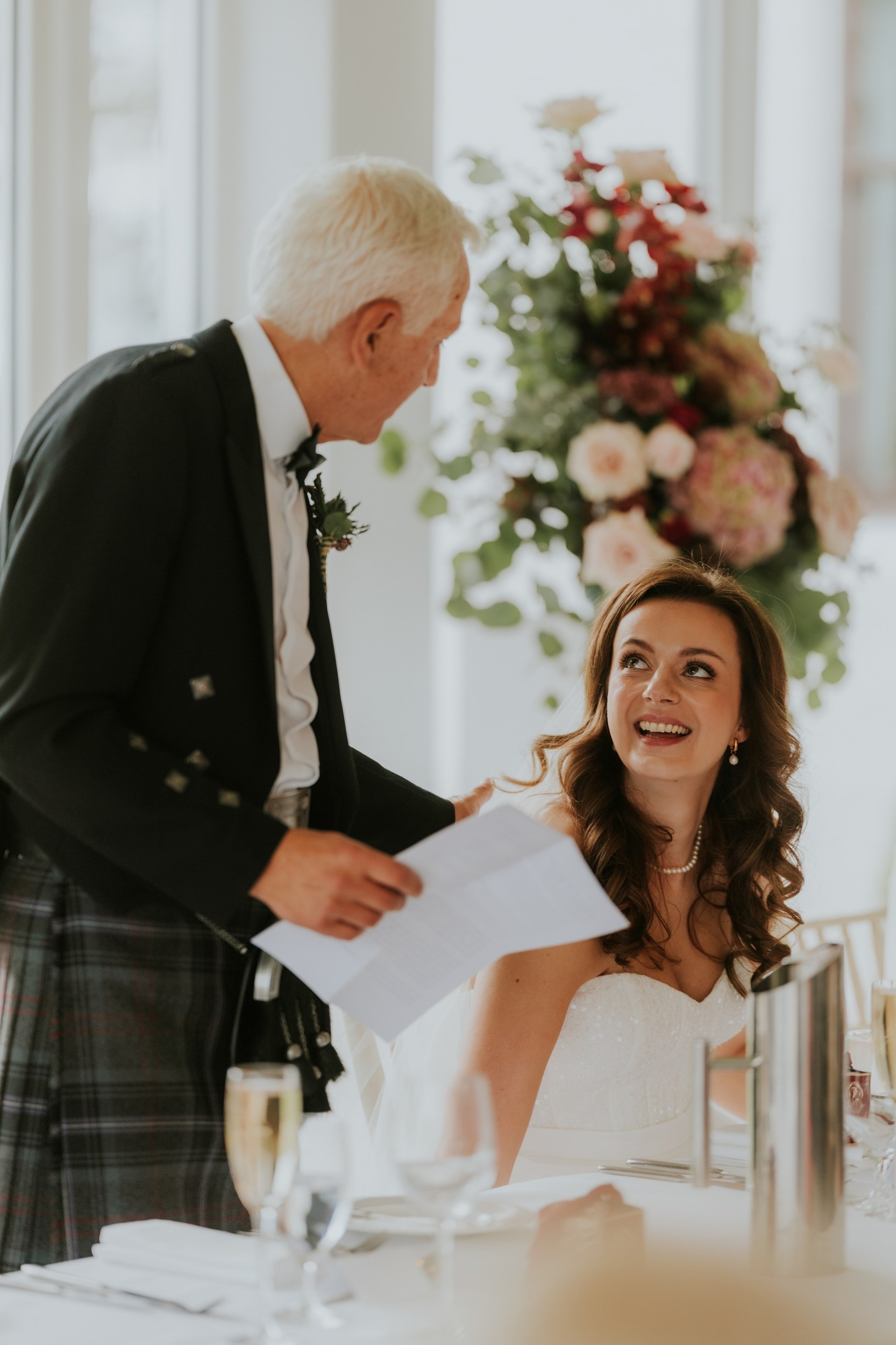 Emma smiles up at a man in a kilt holding speech notes at Lochgreen House Hotel.