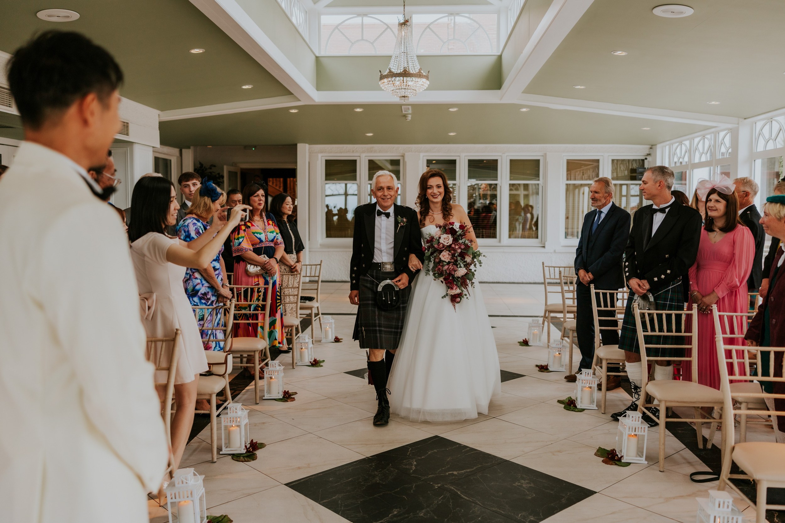 Emma walks down the orangery aisle at Lochgreen House Hotel, holding a cascading bouquet and linking arms with a man in a kilt.