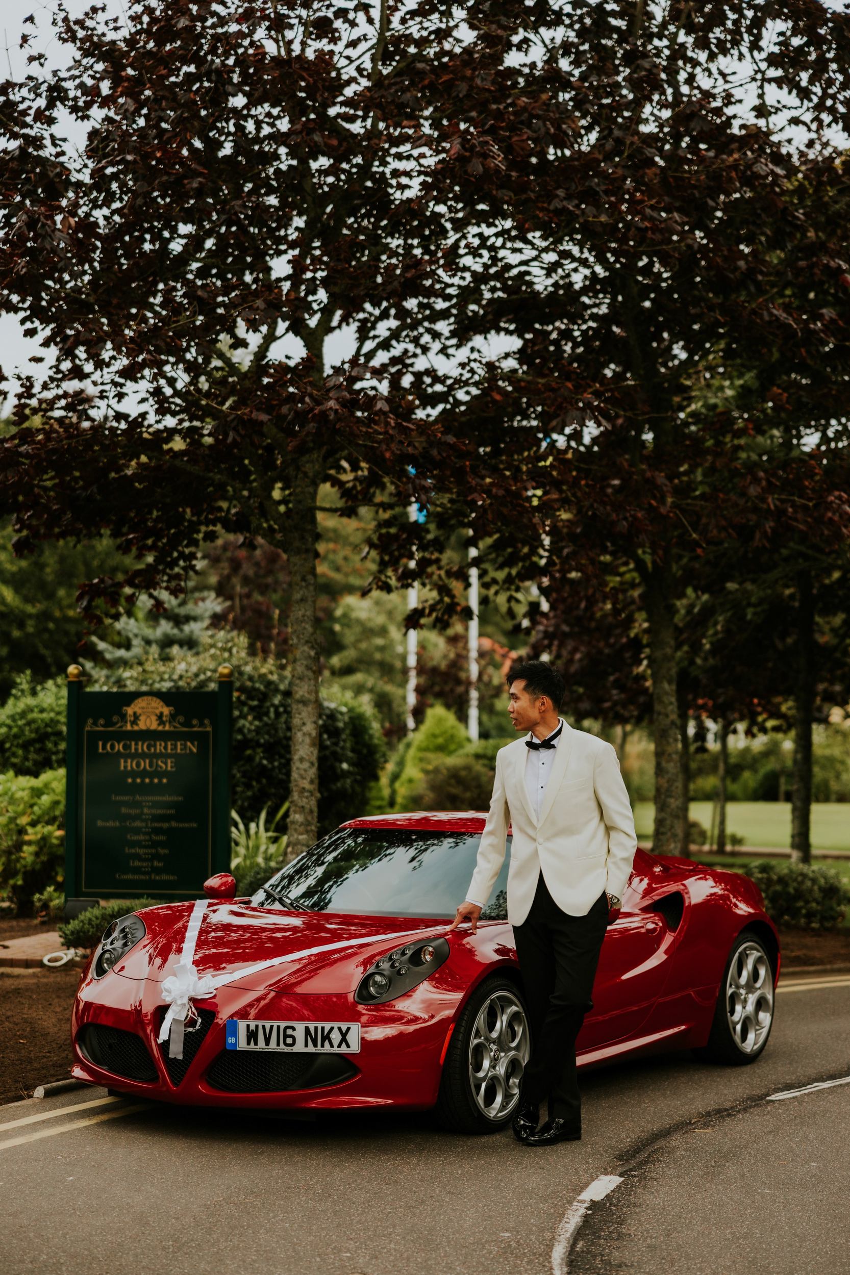 Gordon in a white dinner jacket beside a ribbon-decorated red sports car outside Lochgreen House Hotel.