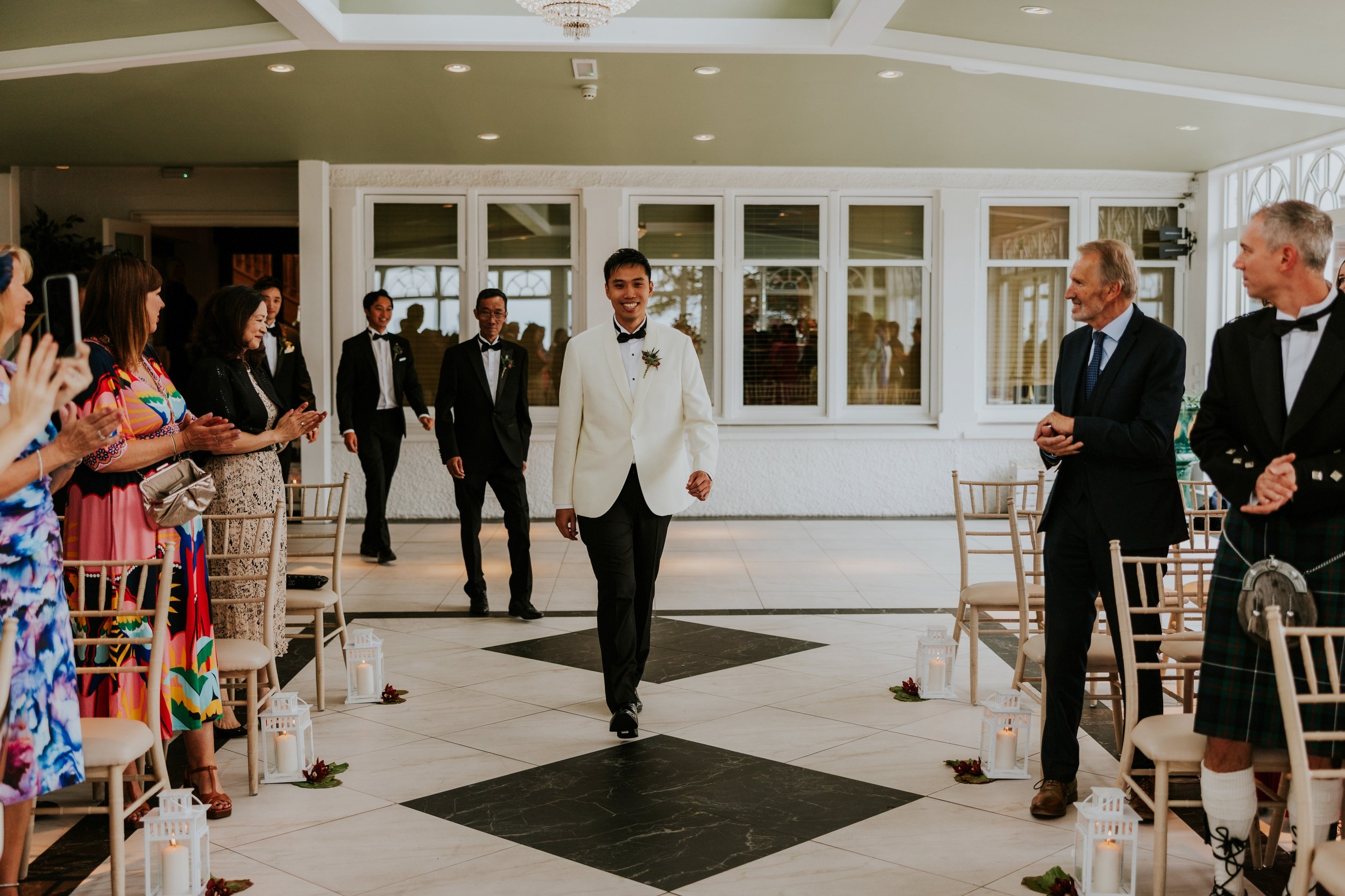 Gordon walks down the aisle in a white dinner jacket as guests applaud in the orangery at Lochgreen House Hotel.