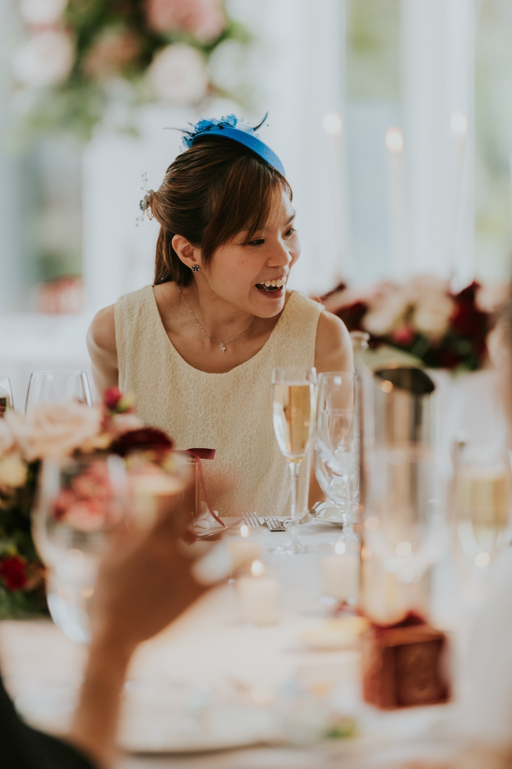 Guest in a blue fascinator smiling at a flower-decorated reception table at Lochgreen House Hotel.
