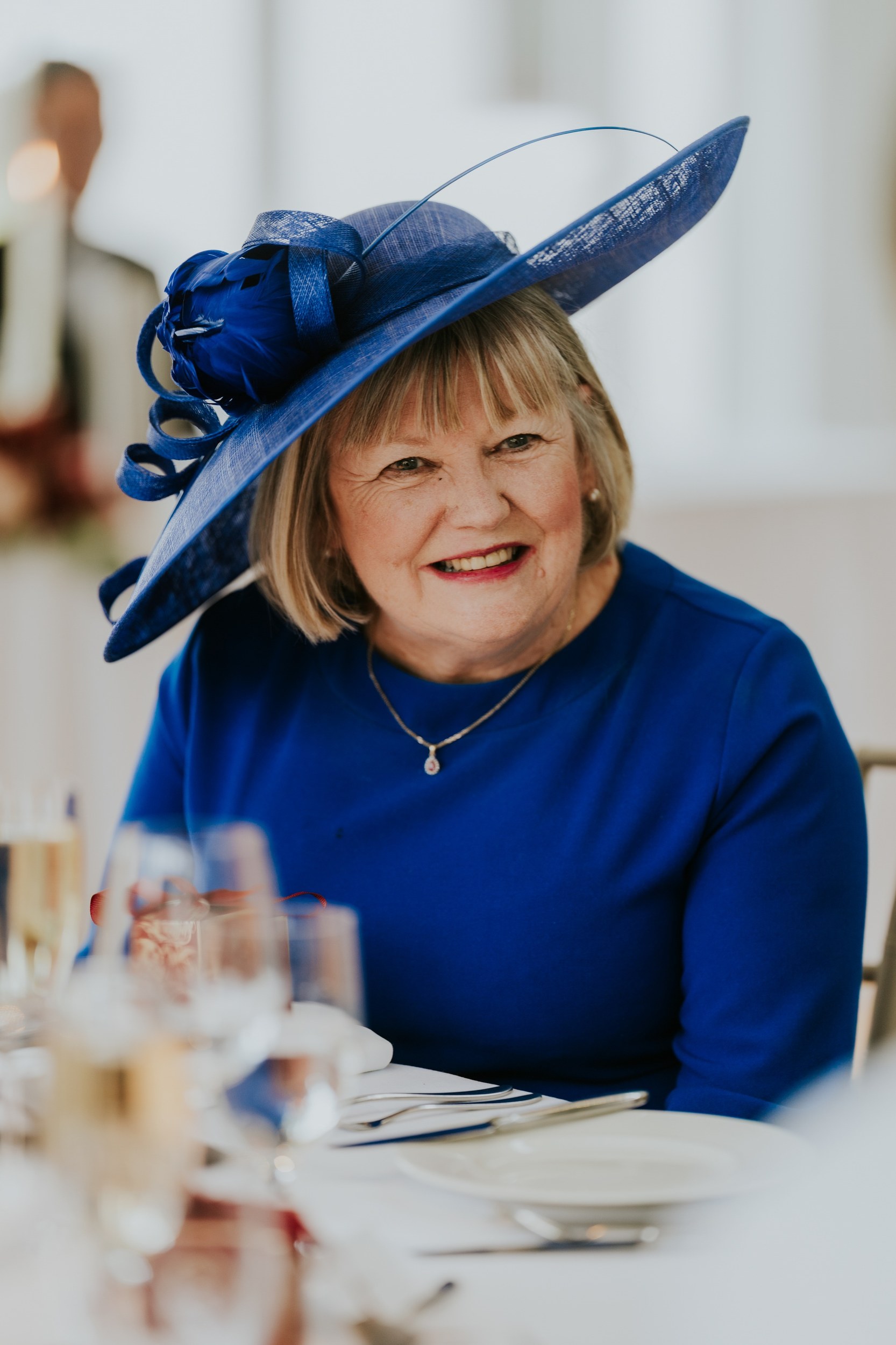 Smiling wedding guest wearing a royal blue outfit and feather-trimmed hat at a reception table.
