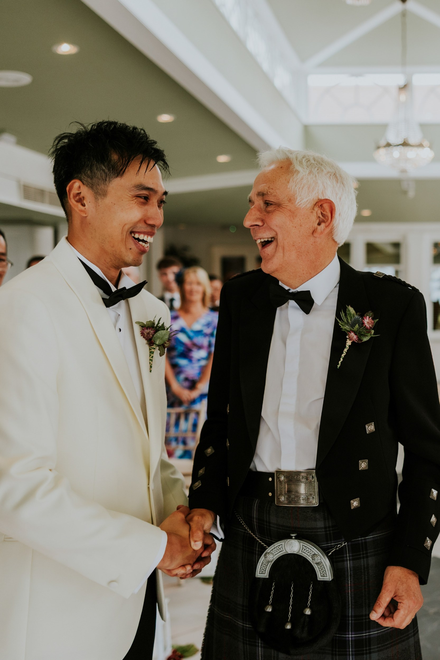 Two men in a white dinner jacket and Highland dress smile and shake hands at Lochgreen House Hotel.