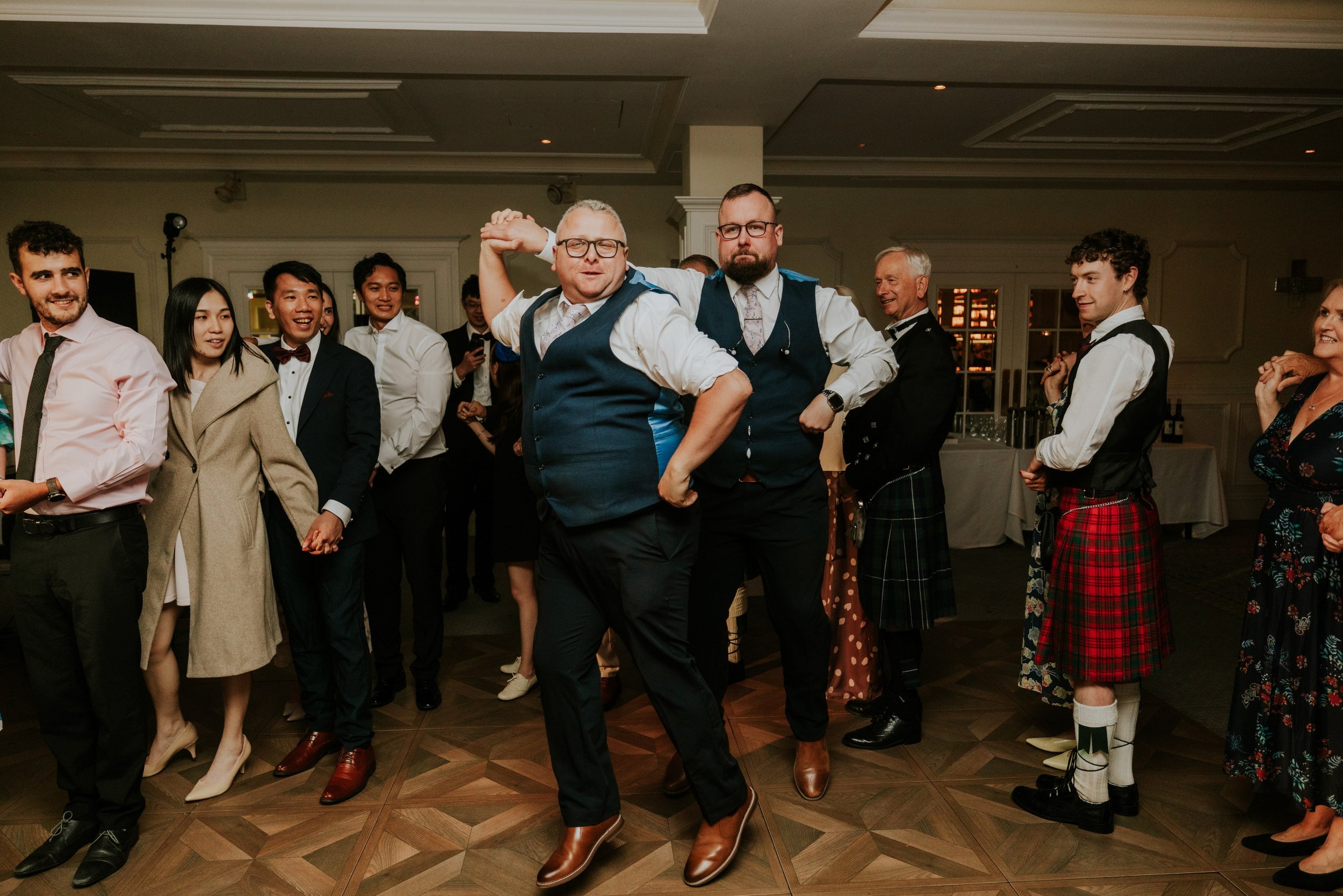 Two men in blue waistcoats dancing with joined hands as wedding guests watch at Lochgreen House Hotel.