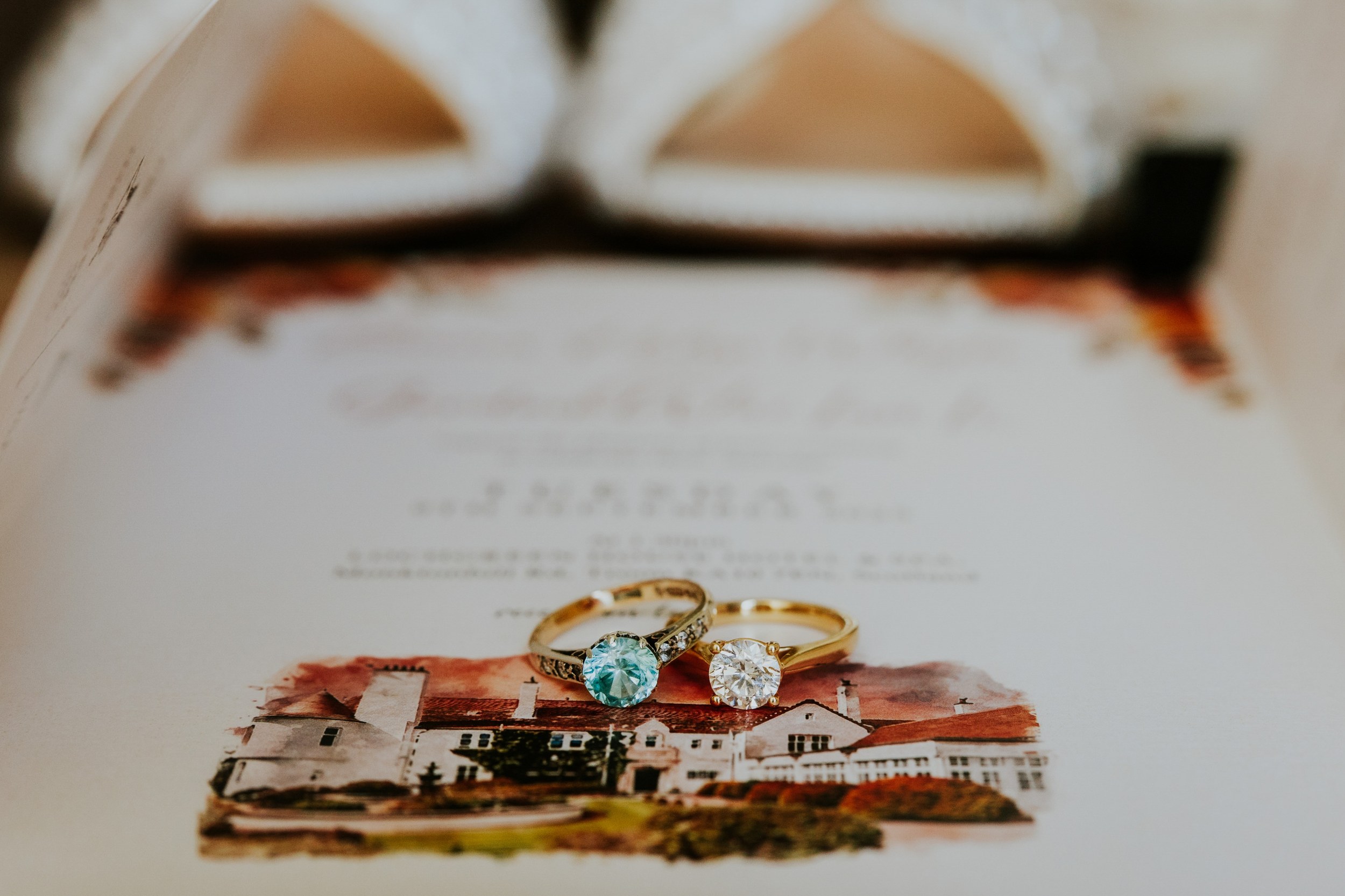 Two rings with blue and clear stones on wedding stationery illustrated with Lochgreen House Hotel, with white shoes behind.