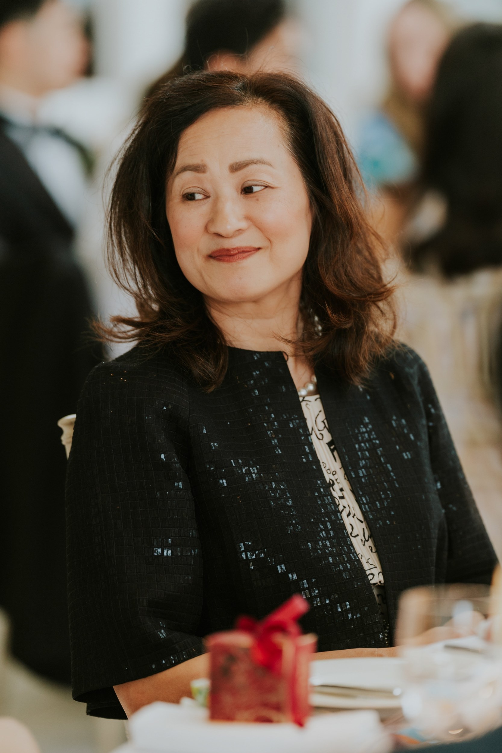 Wedding guest in a dark jacket smiling at a laid table, with a red favour box and glassware in front of her.