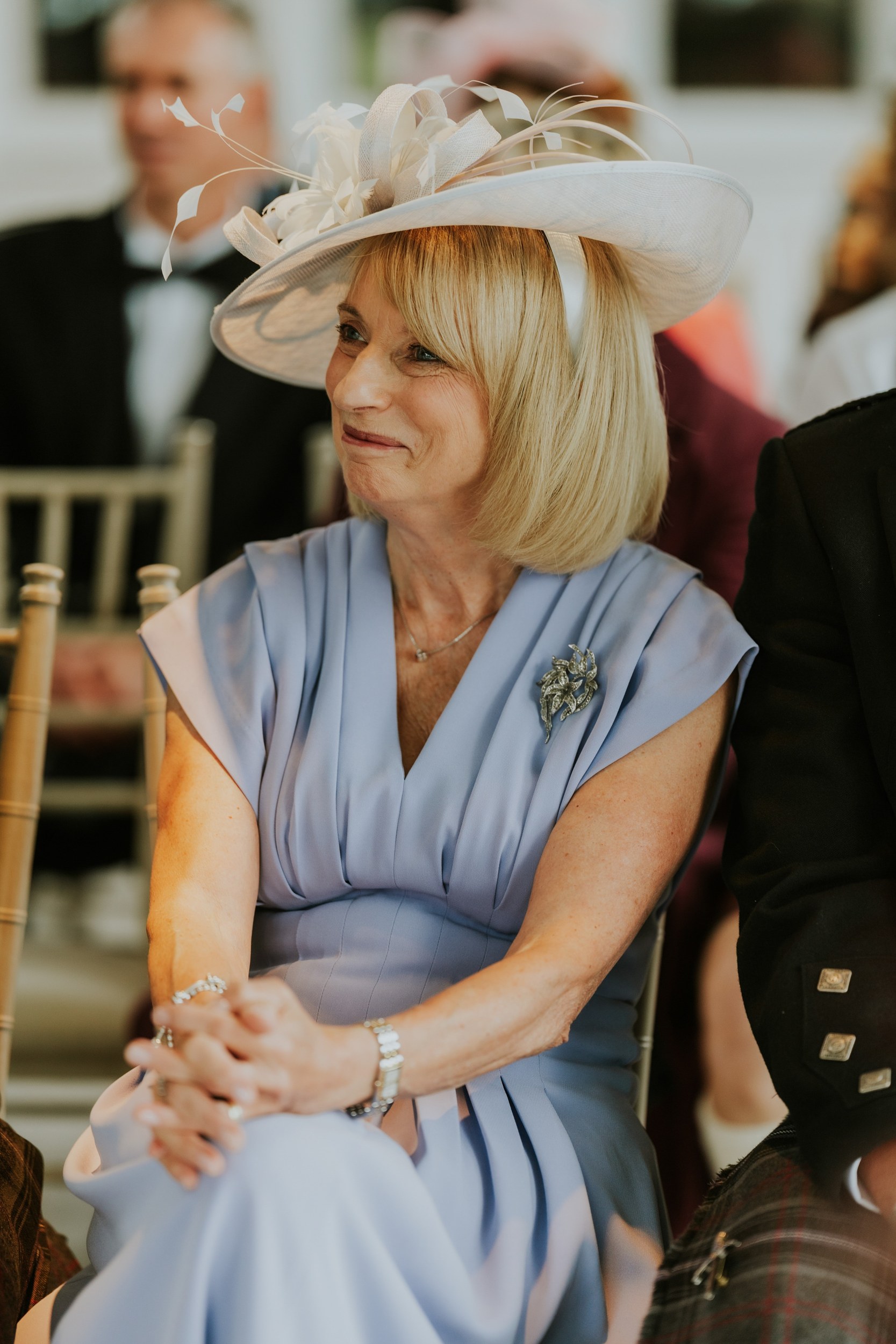 Wedding guest in a pale blue dress and cream hat, seated and smiling among other guests at Lochgreen House Hotel.