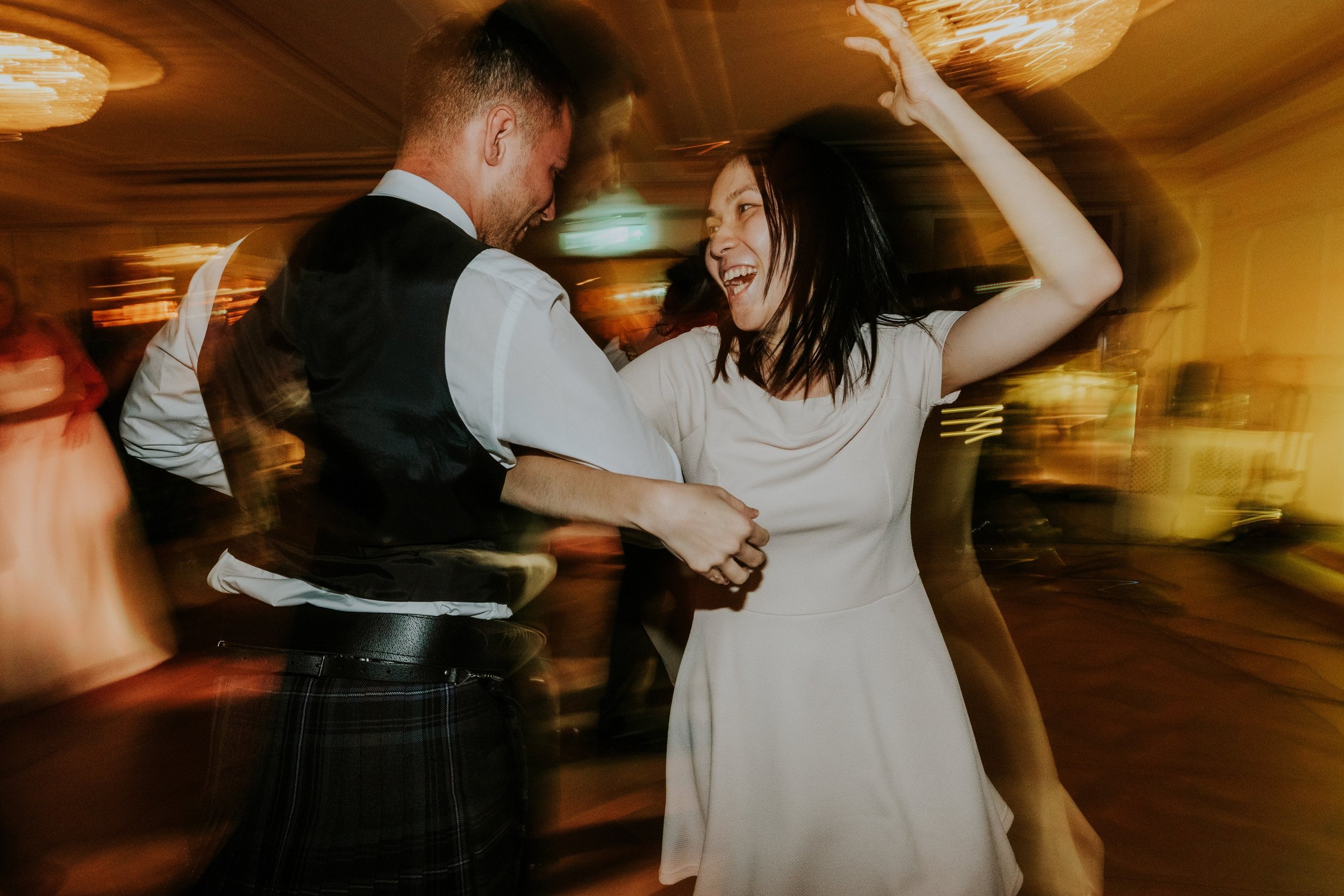 Woman in a white dress raises her arm while dancing with a man in a tartan kilt at Lochgreen House Hotel.