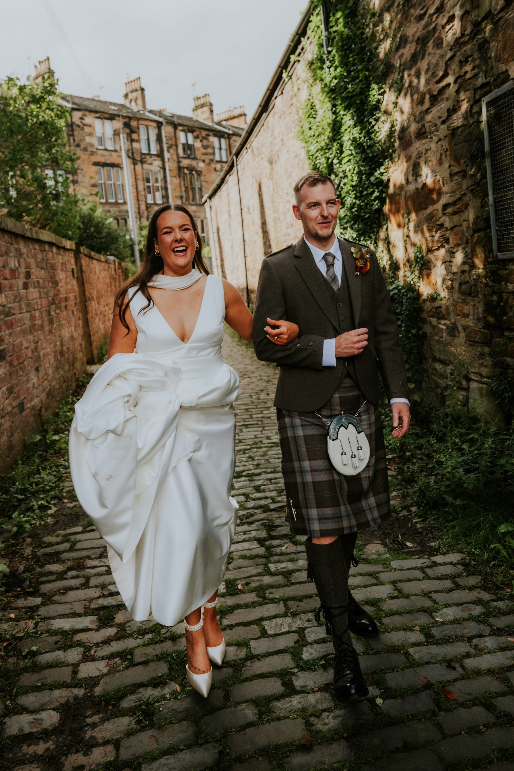 Bride and groom arm-in-arm walking down a cobbled alley by stone walls, bride laughing and groom smiling.