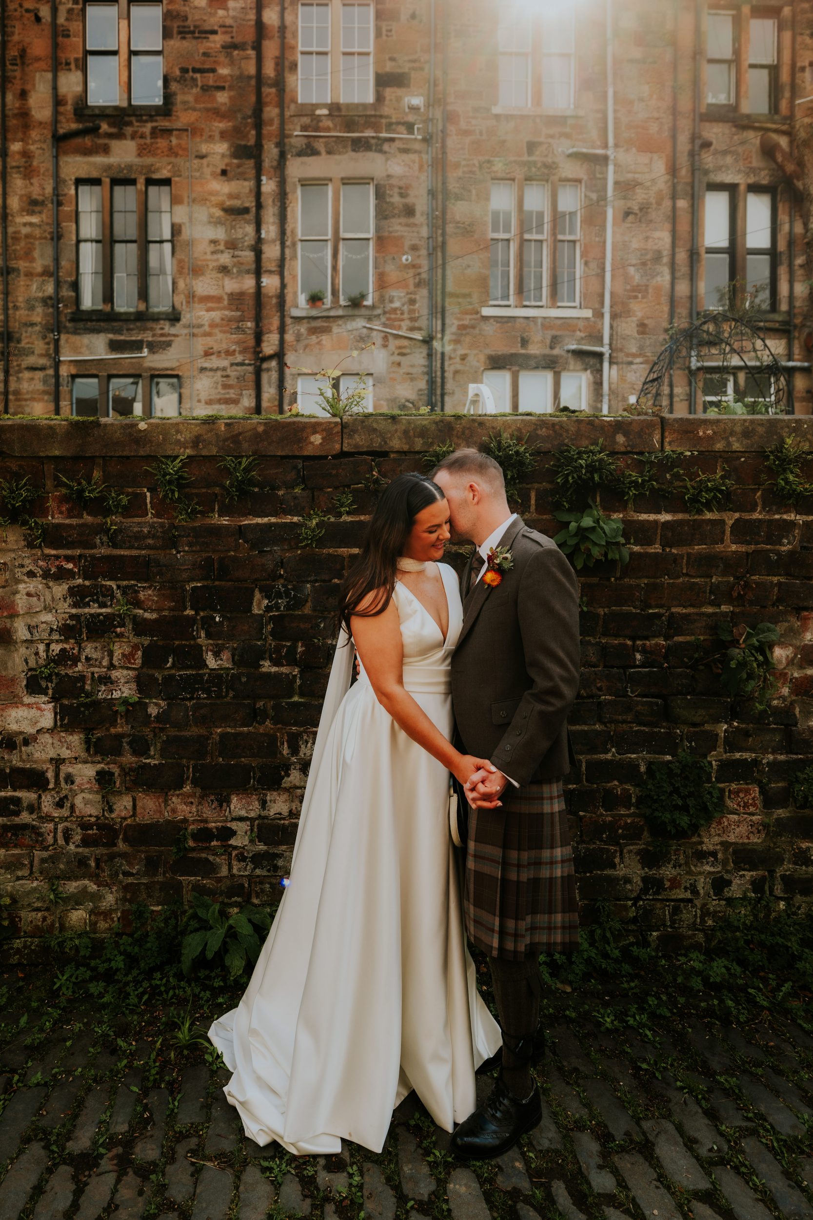 Bride and groom hold hands and touch foreheads in front of a mossy brick wall; groom in kilt, bride in a white sleeveless dress.