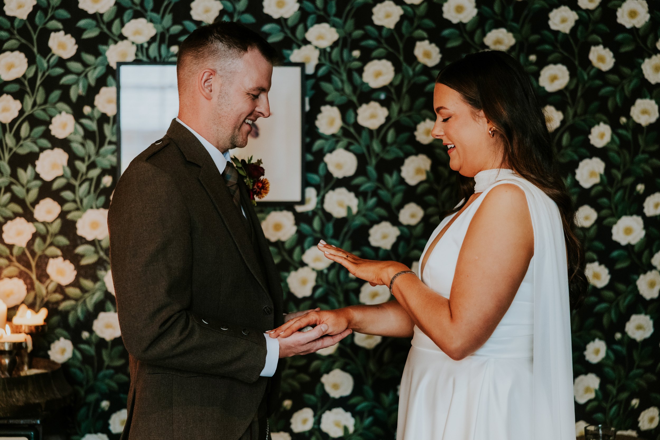 Bride and groom smiling as they exchange rings against dark floral wallpaper.