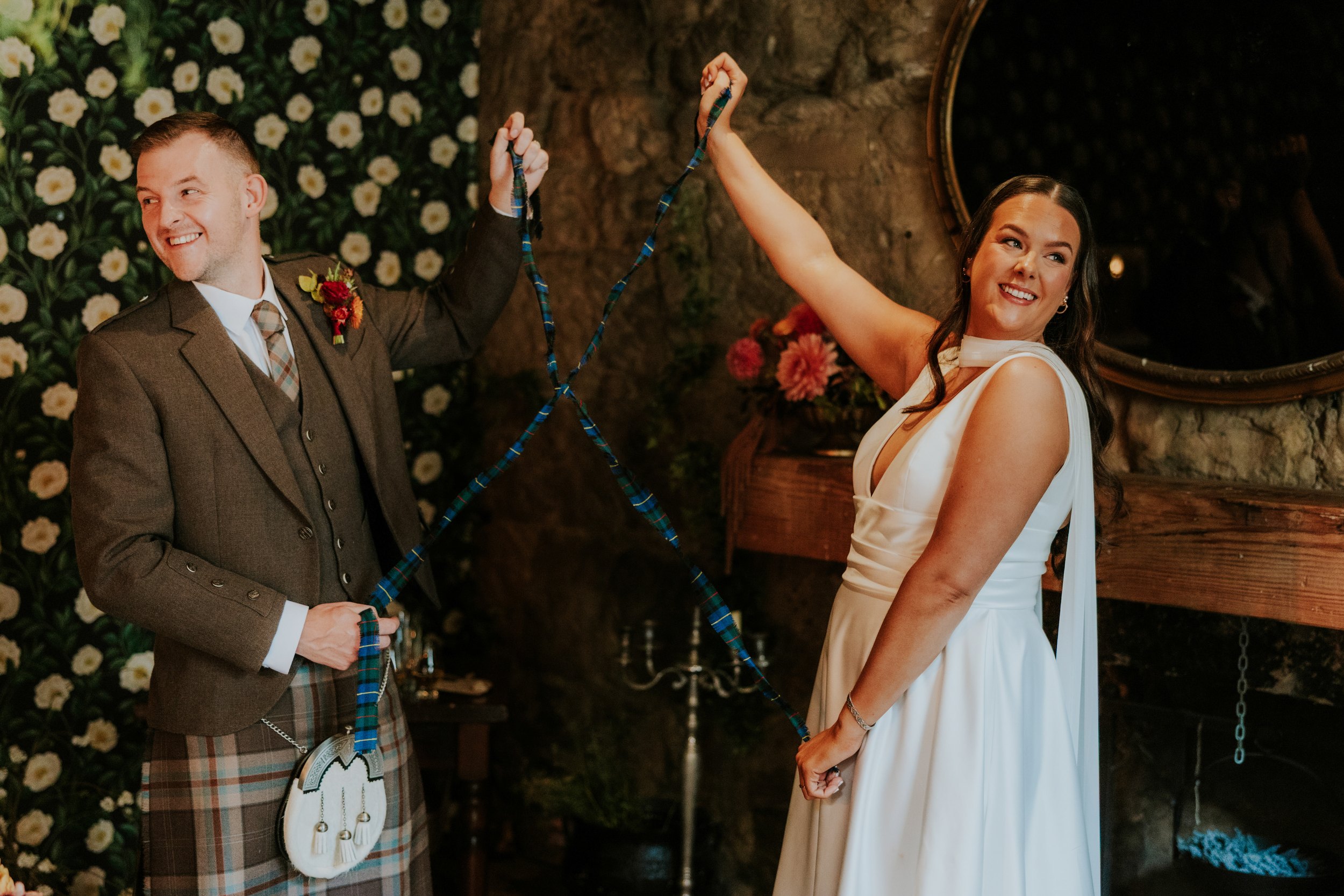 Bride and groom smiling while holding tartan handfasting cords inside The Bothy.