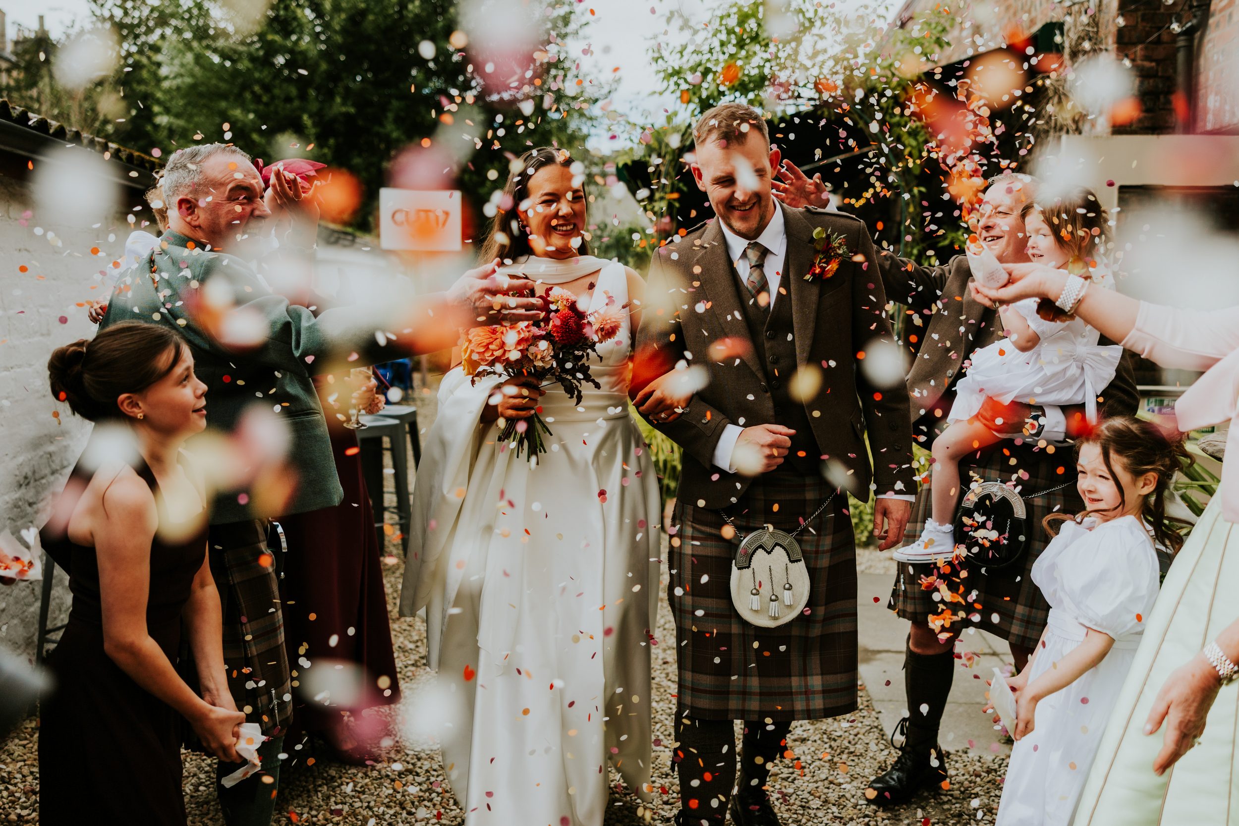 Bride and groom walk through colourful confetti tossed by guests outside; bride holds bouquet, groom in kilt with sporran.