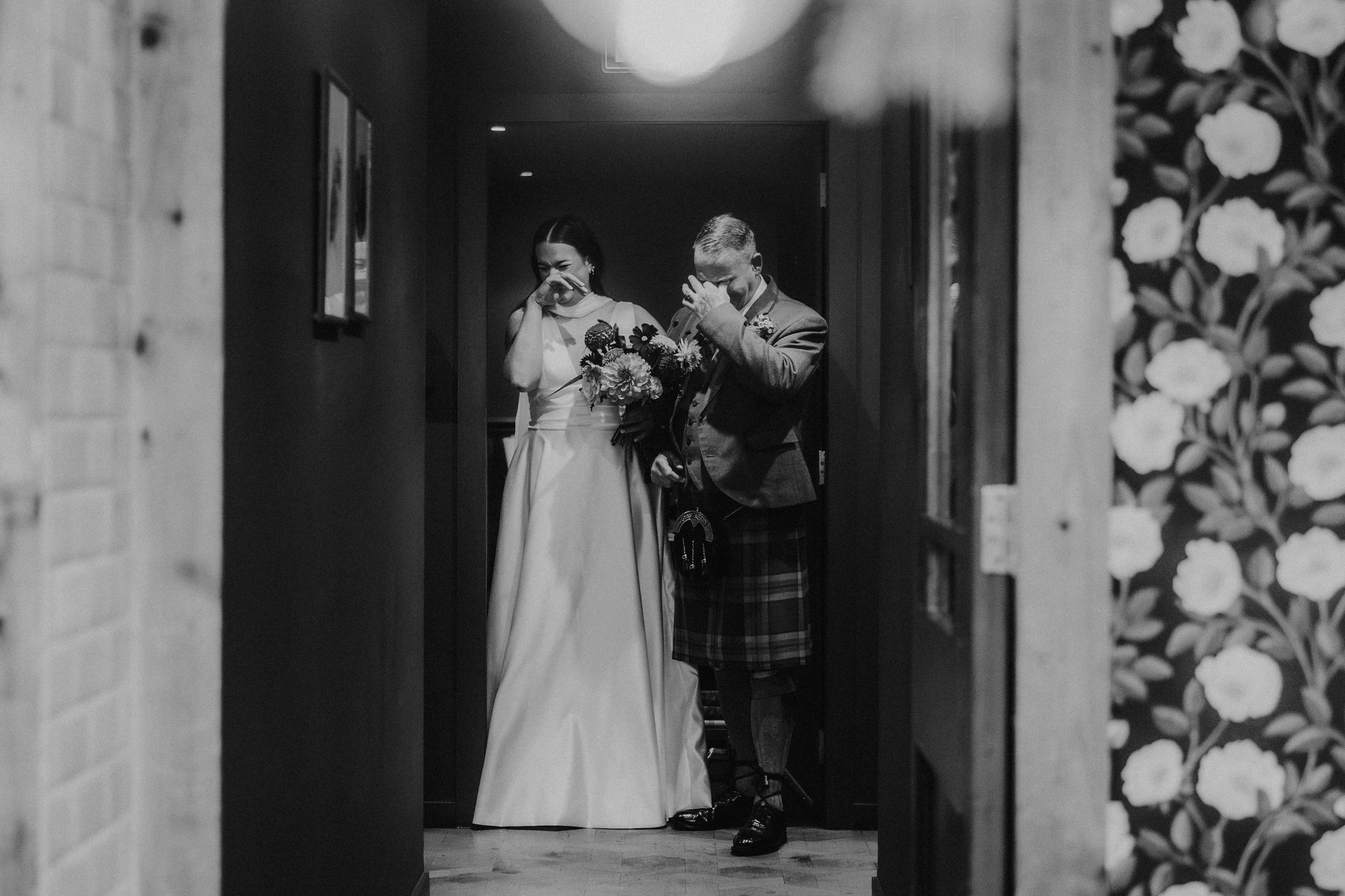 Bride in a sleeveless gown and groom in a kilt stand in a doorway, both wiping away tears while the bride holds her bouquet.