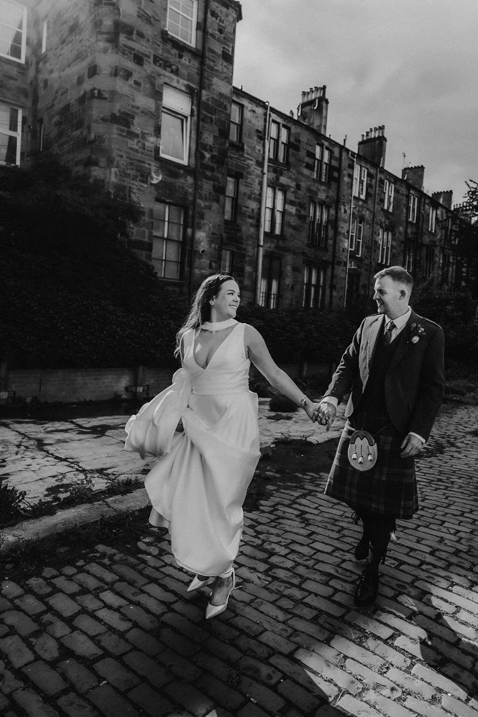 Bride in a wedding gown and groom in a kilt holding hands and walking along a cobbled street beside sandstone tenement buildings.