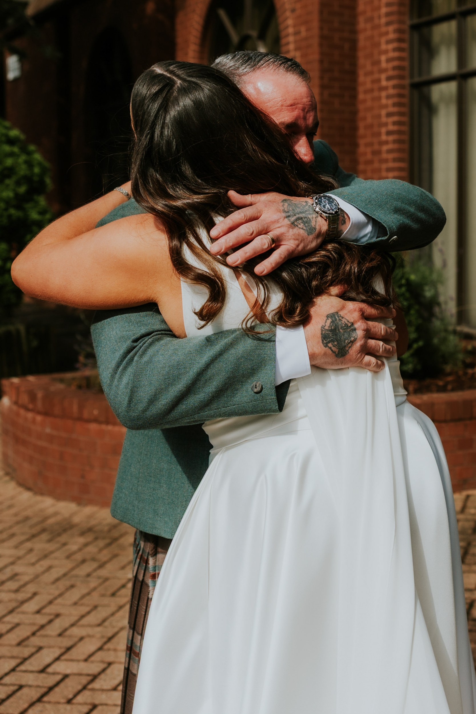 Bride in a white dress embraces an older man in a green jacket and kilt outside a brick building.