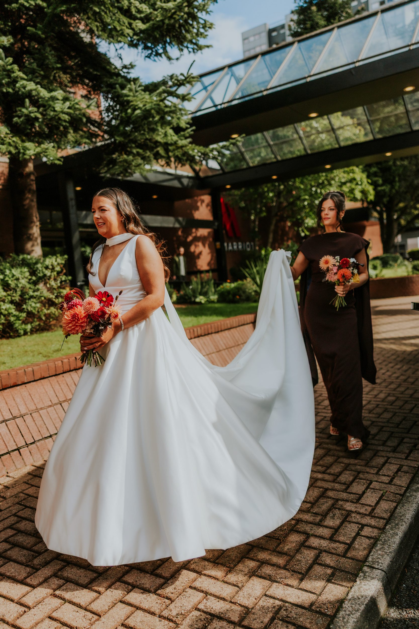 Bride in a white gown walking outside with bouquet while a bridesmaid carries her long train by a hotel entrance.