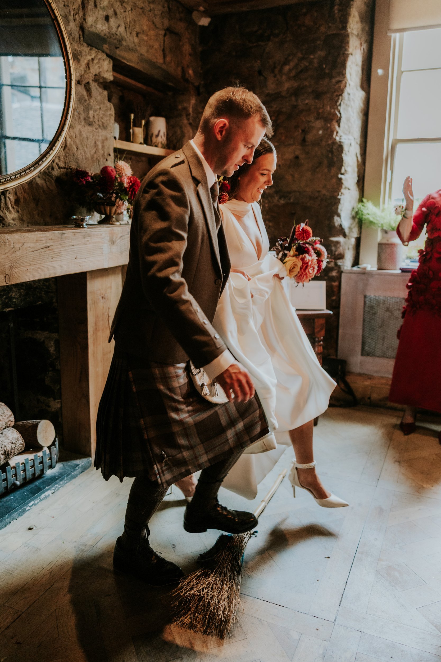 Bride in white and groom in a kilt step together over a broom inside a stone-walled room, bride holding a bouquet.