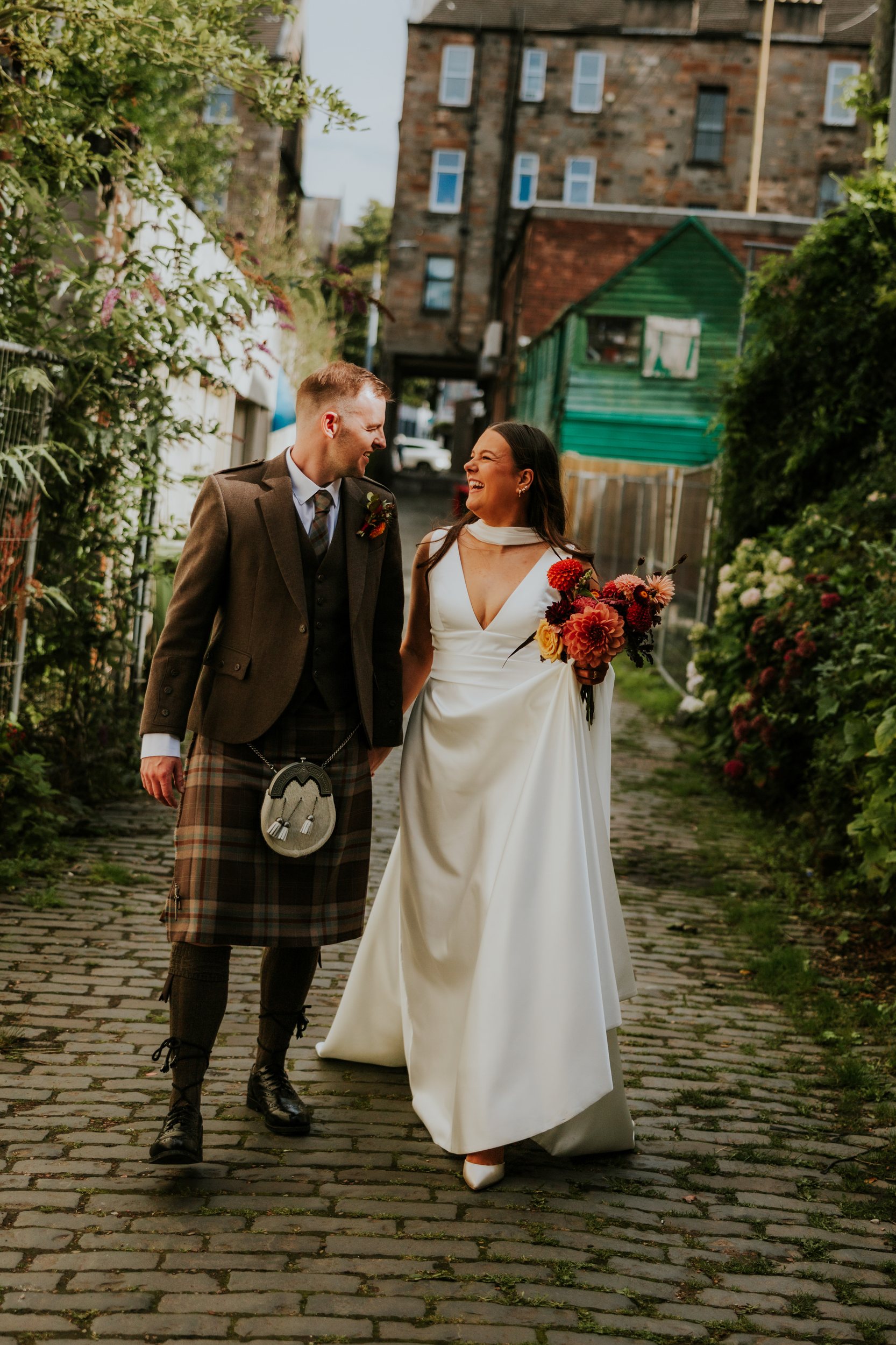 Bride in white dress and groom in kilt laughing as they walk along a cobbled lane, bride holding a bouquet.
