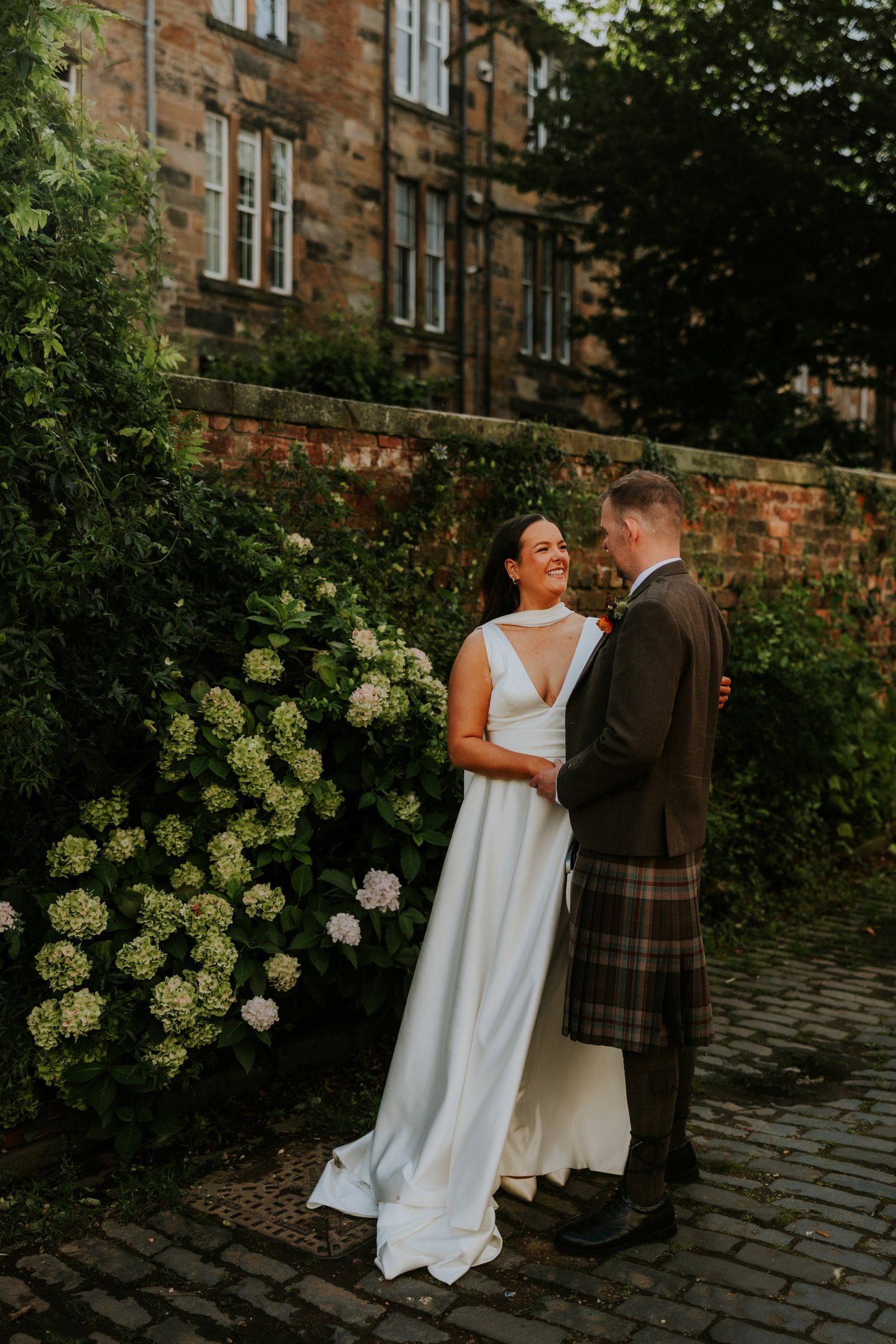 Bride in white dress and groom in kilt hold hands and smile by a brick wall and hydrangeas.