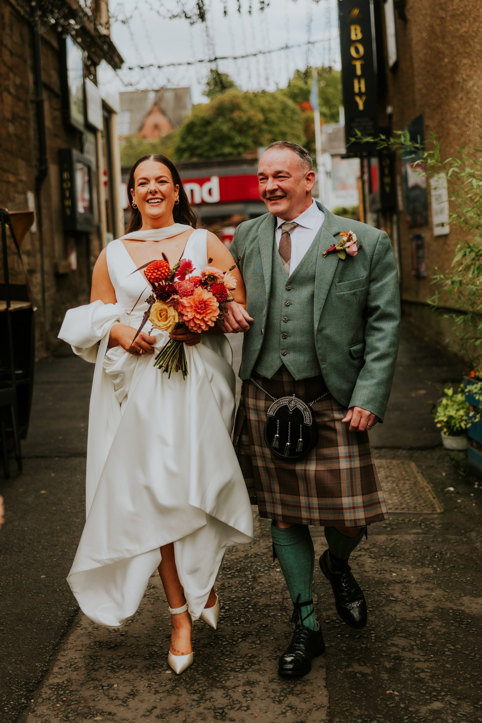 Bride in white dress with bouquet walking arm-in-arm with a man in a kilt outside The Bothy, both smiling.