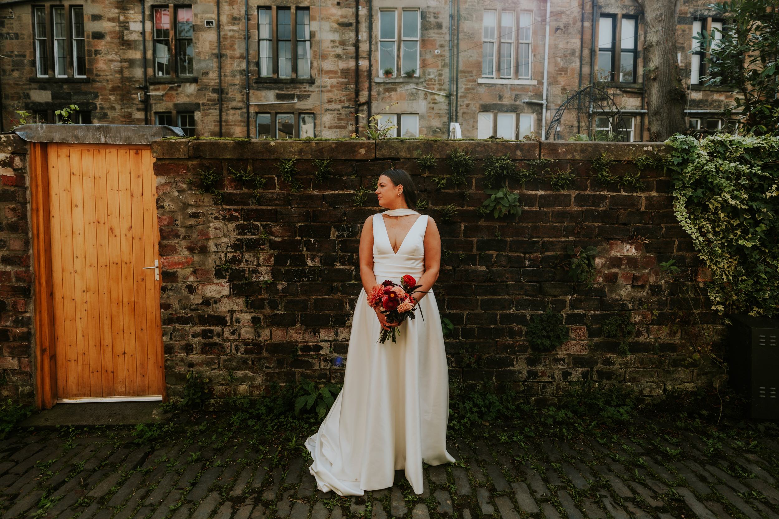 Bride in white gown holding bouquet, standing against a mossy brick wall beside a wooden gate.