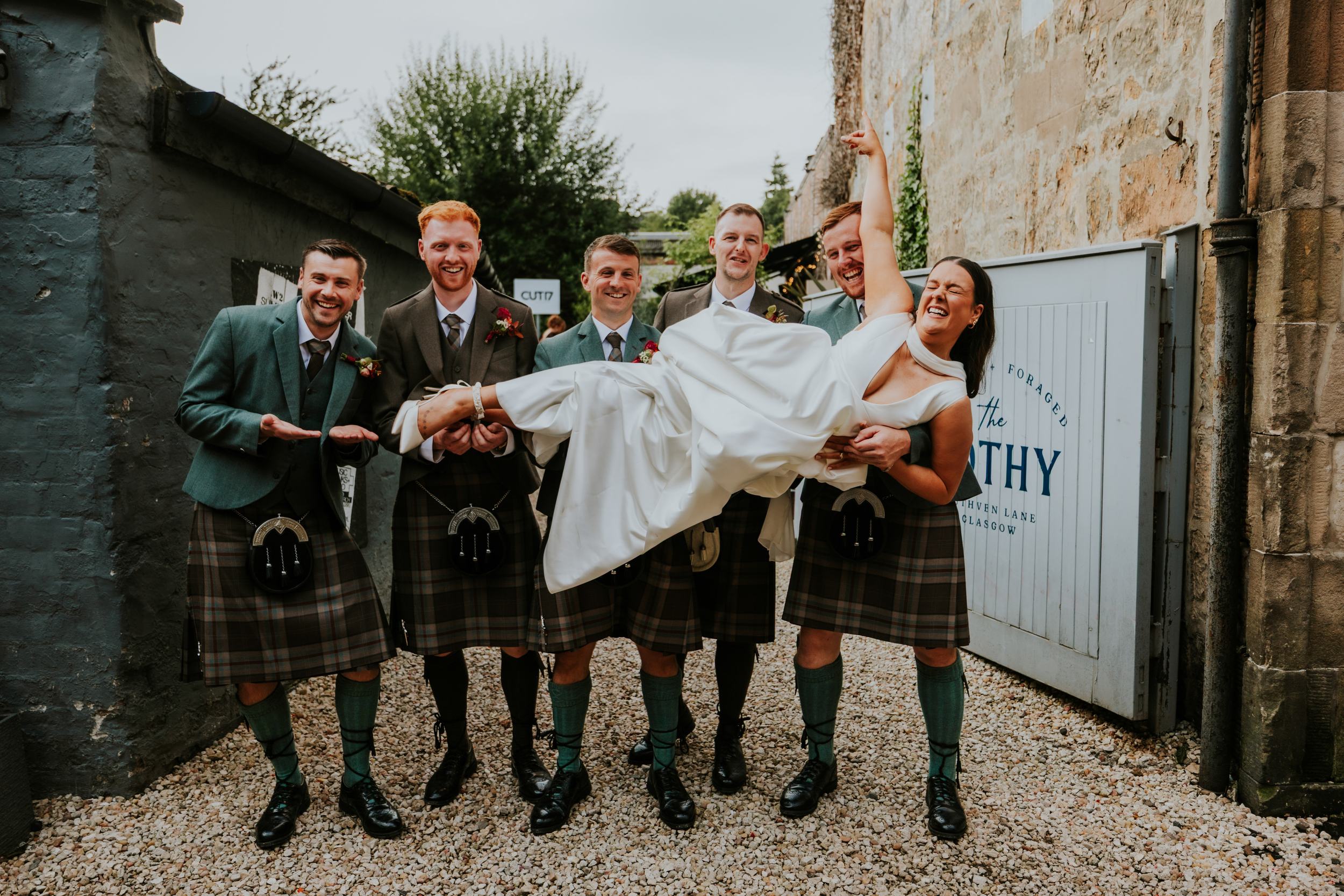Bride laughing as six kilted groomsmen hold her aloft outside The Bothy courtyard.