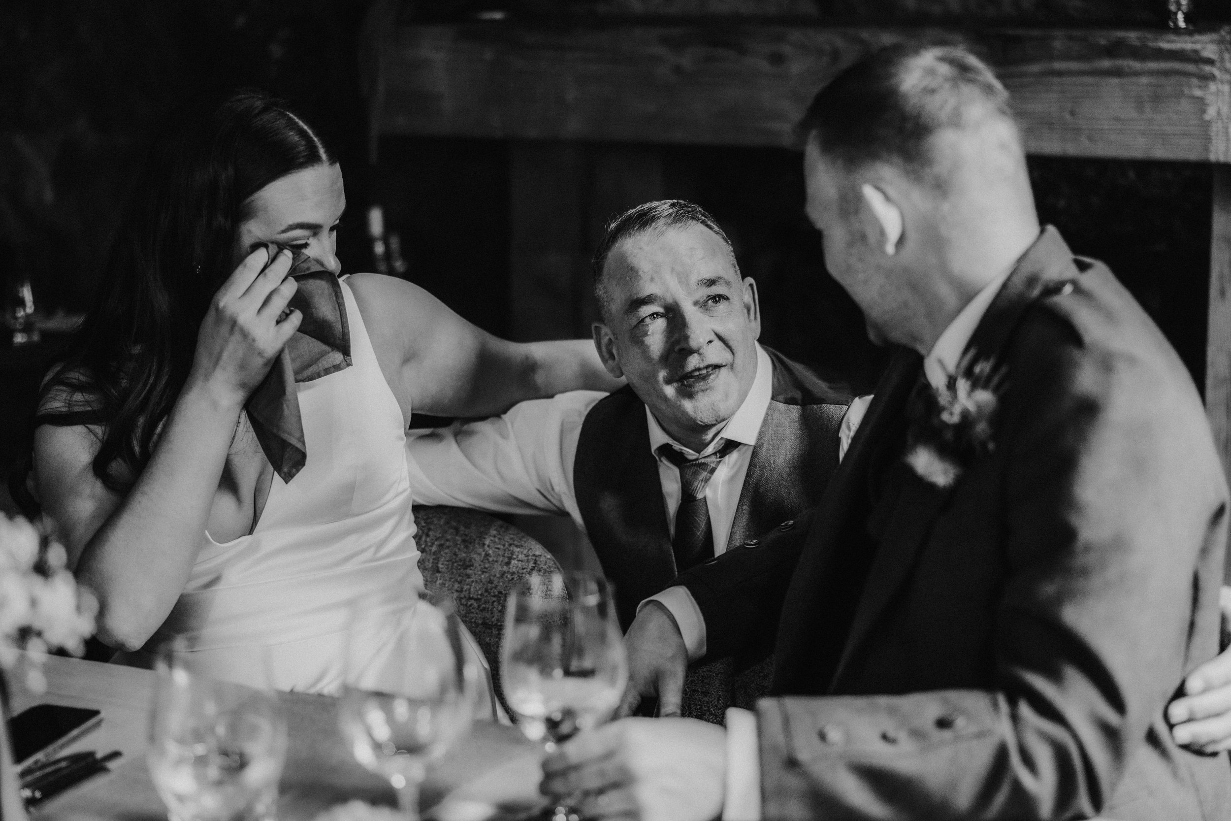 Bride wiping tears with a napkin as two men sit close by and talk at a table.