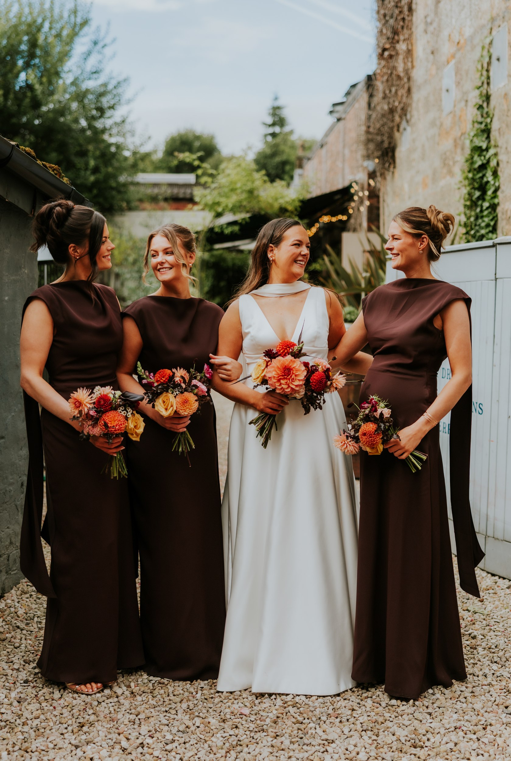 Bride with three bridesmaids in dark dresses, holding bouquets and smiling together in a courtyard.