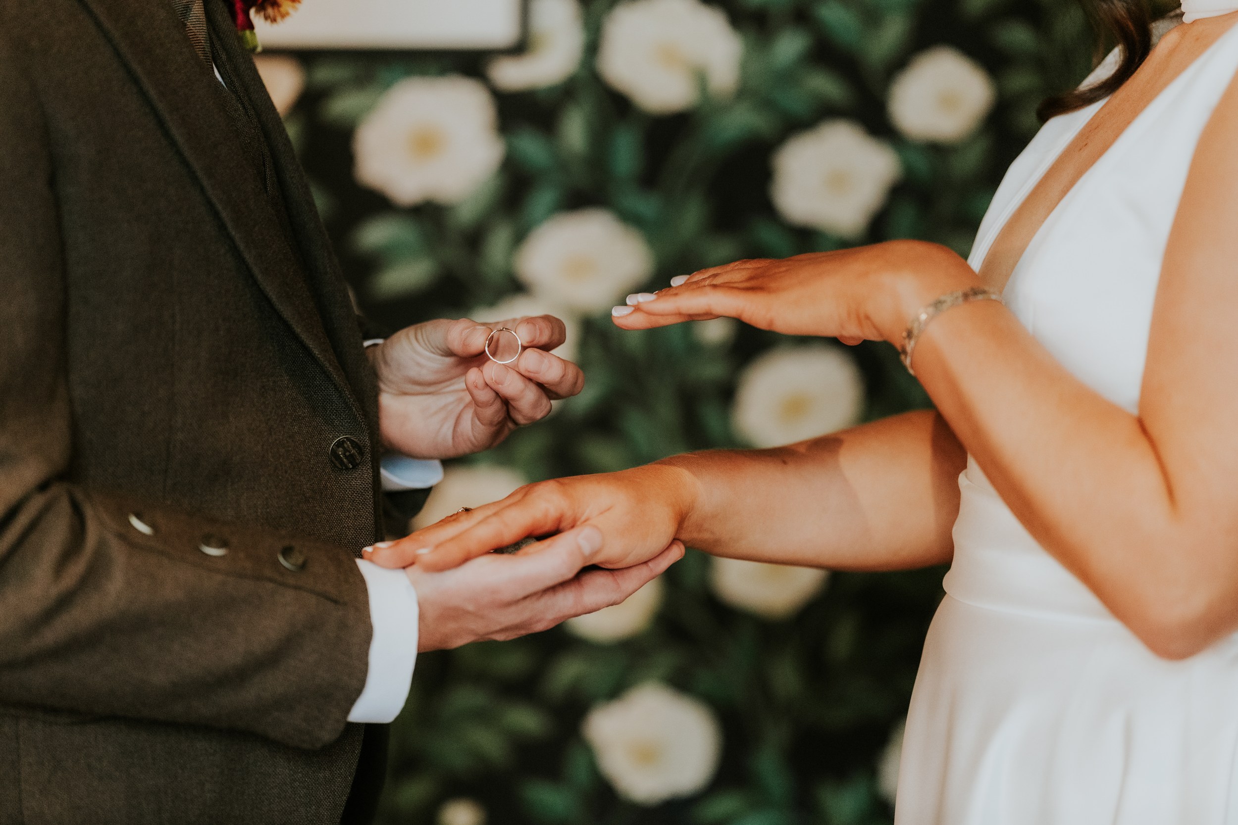 Close-up of bride and groom exchanging rings; groom holds ring as bride's hands are outstretched against a floral backdrop.
