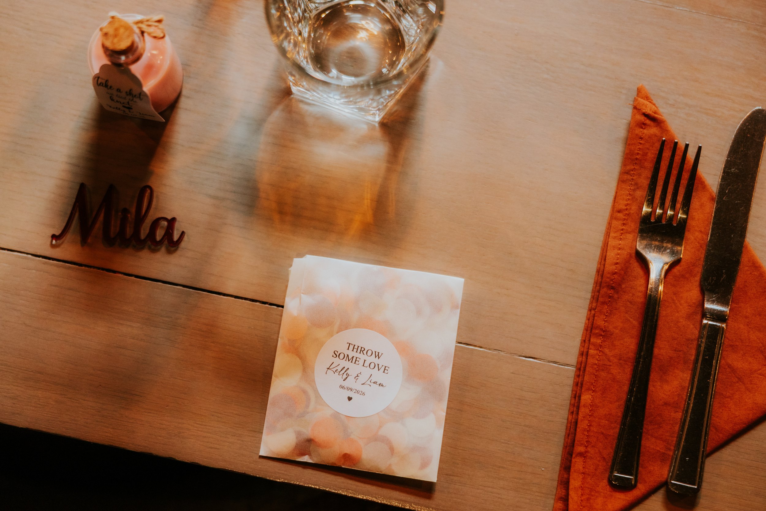 Close-up of wedding place setting with knife and fork on orange napkin, glass, confetti bag labelled Kelly & Liam and wooden name 'Mila'.