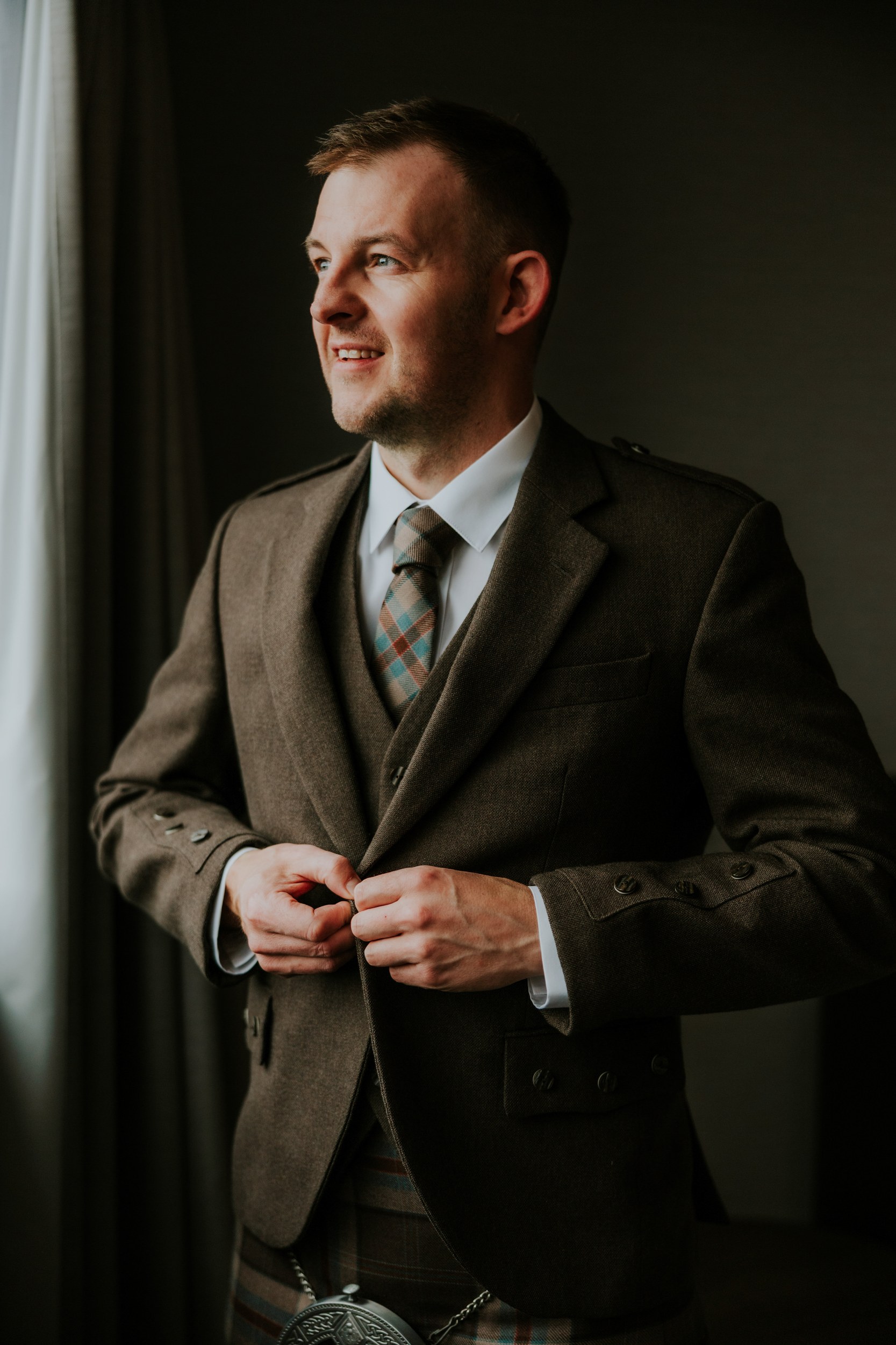 Groom in brown tweed jacket and tartan kilt buttoning his jacket by a window, looking out.