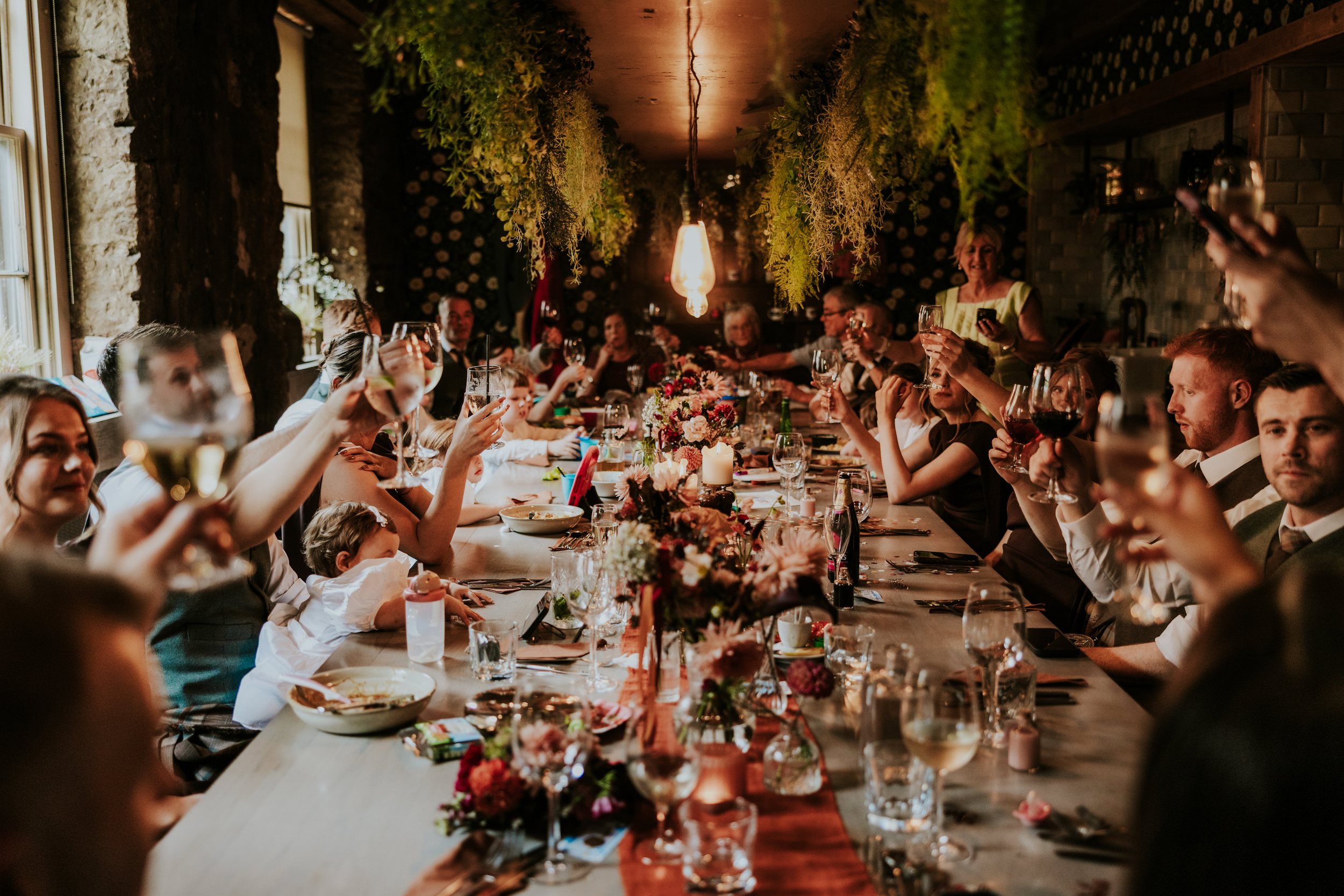 Guests at a long decorated wedding table raise glasses in a toast under hanging greenery and warm light.