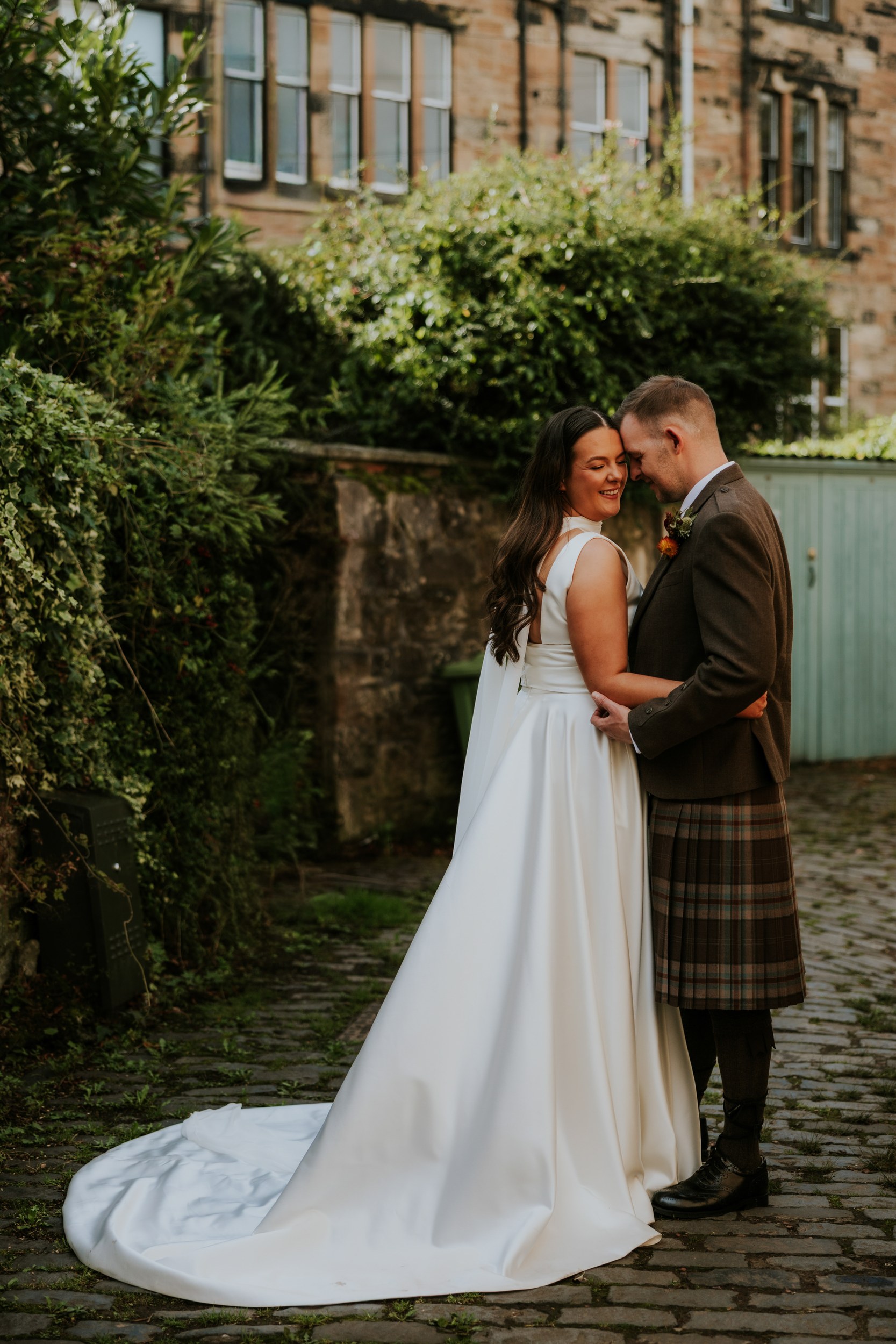 Kelly and Liam forehead-to-forehead embrace in wedding attire on a cobbled path beside a stone wall and greenery.