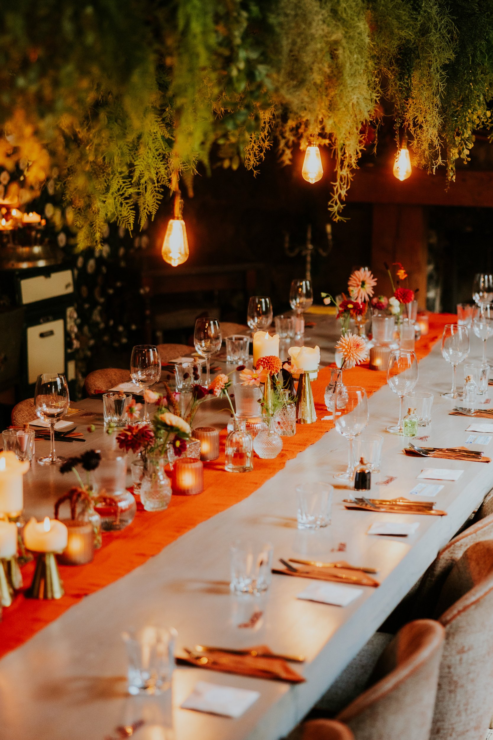 Long wedding reception table with glasses, candles and small flower vases beneath hanging greenery and warm filament bulbs.