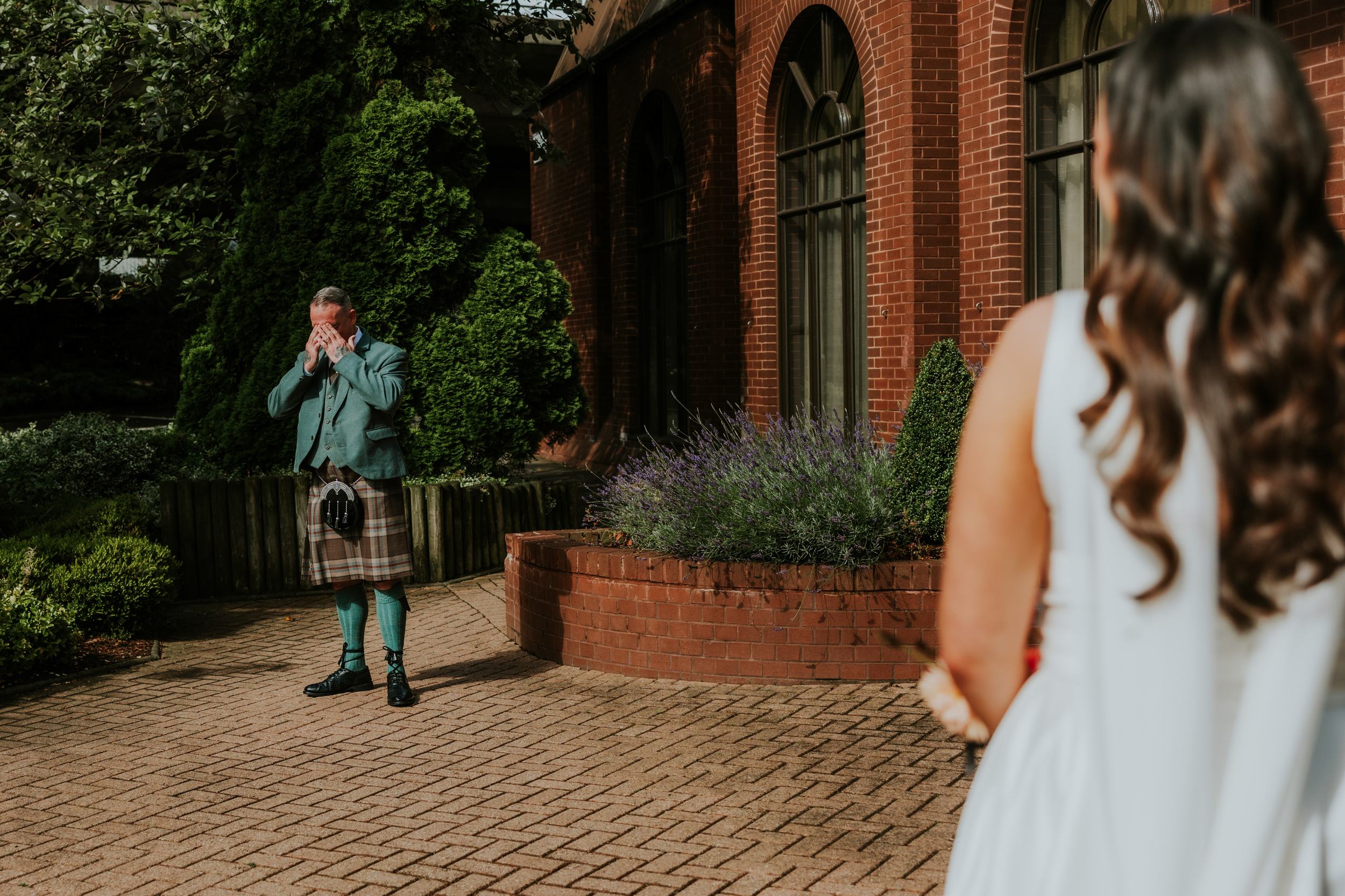 Man in kilt wiping his face as a bride approaches outside a red-brick building.